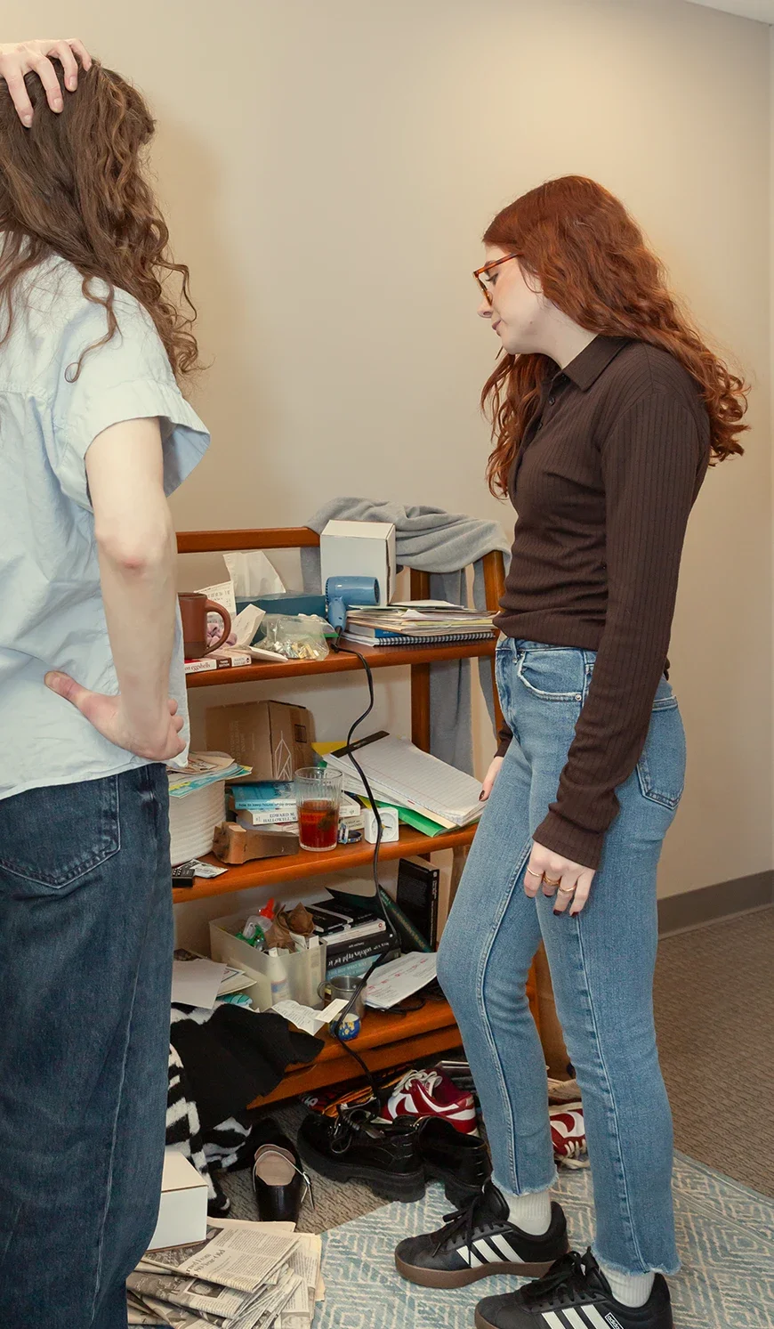 Two women with red hair in casual clothing converse in a cluttered room with shelves full of books, papers, and miscellaneous items on the floor.