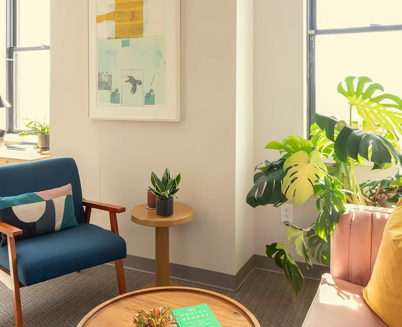 Green Fig's therapy & coaching office in Philadelphia, with a blue armchair & graphic pillow, a cozy pink velvet sofa & yellow plush pillow, a large green monstera plant, a wooden coffee table with a succulent & books, & framed artwork on the wall.