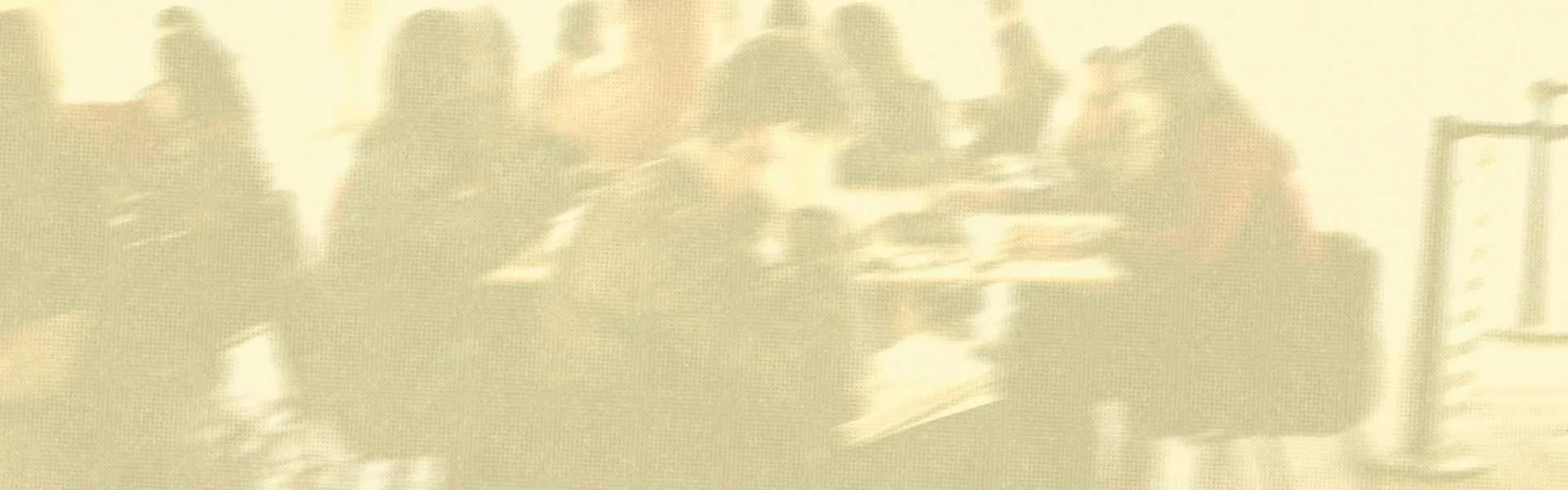 People sitting at tables in a room, possibly a cafeteria or conference room, with some looking at their phones or talking.