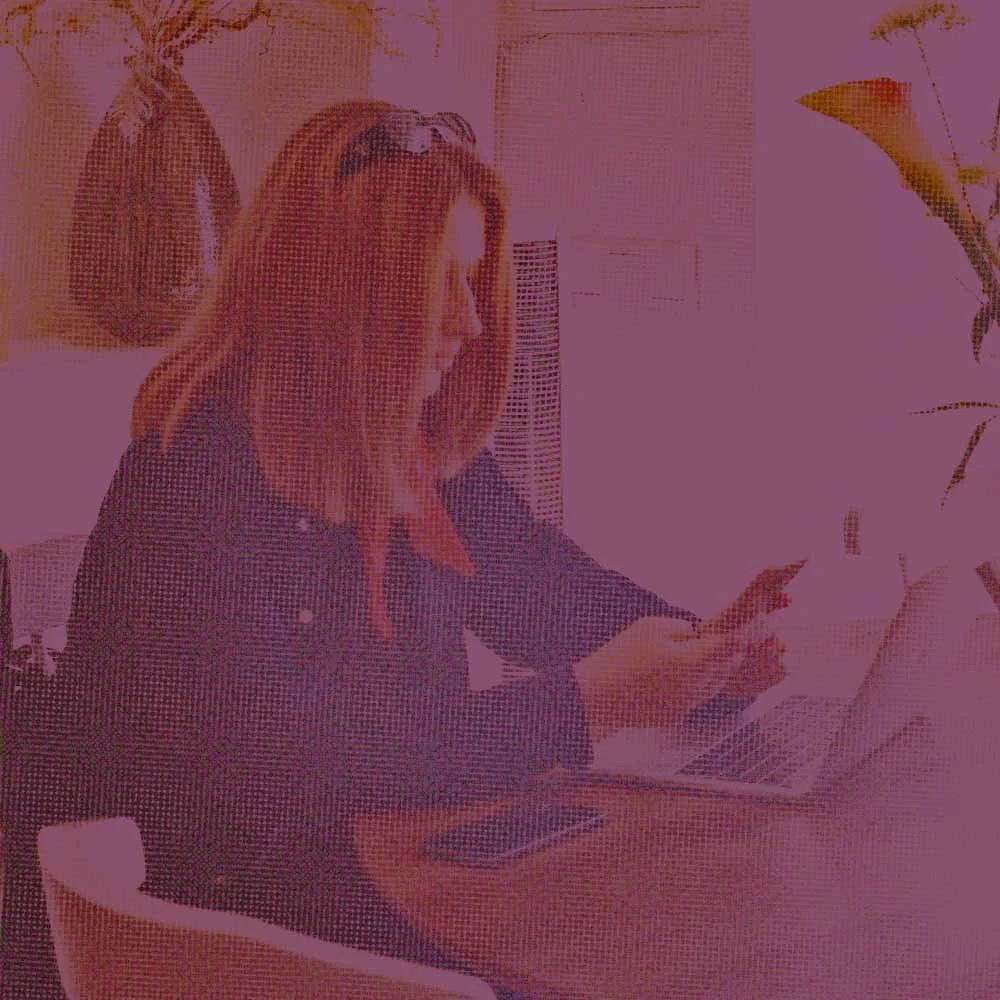 A woman with red hair sitting at a desk, working on a laptop and using her phone in an indoor environment.