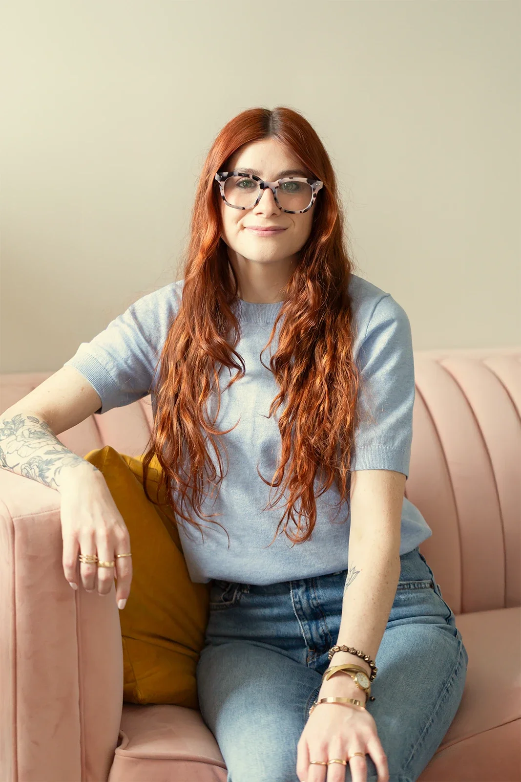Molly Marcus, therapist & coach specializing in DBT & Decluttering, with long red hair, glasses, and tattoos, wearing a gray t-shirt & jeans and sitting on a pink sofa with a yellow pillow in her Philadelphia office.