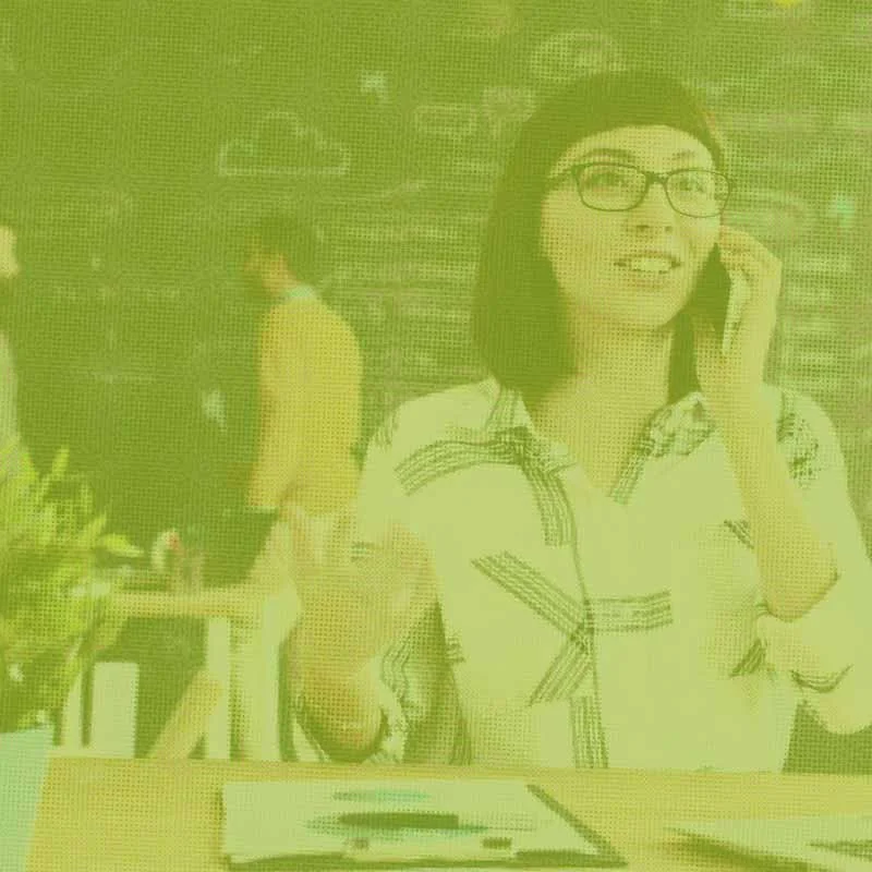 A young woman with glasses talking on her cellphone at a restaurant table with a notepad and pen in front of her.