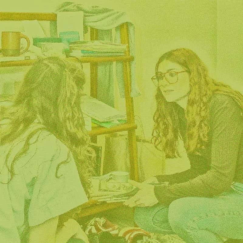 Two women with long curly hair and glasses talking in a cozy room with a bookshelf filled with books and a cup on a table.
