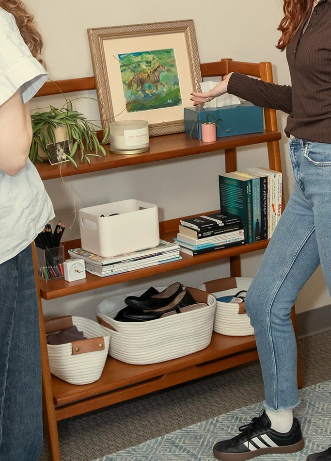 Two women standing next to a wooden bookshelf filled with books, baskets, and decor items, including a framed painting of a horse and a potted plant.
