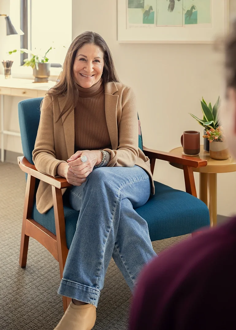 Therapist Dana Marcus, sitting in a blue chair & smiling, in an individual therapy session with a client in her Philadelphia office.