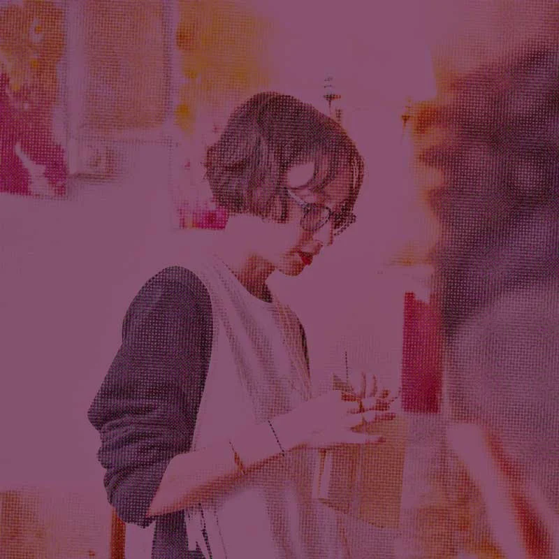 A woman with short dark hair, glasses, and a white shirt with dark sleeves, holding a glass and looking down, in an indoor setting with blurred posters or artwork on the wall behind her.