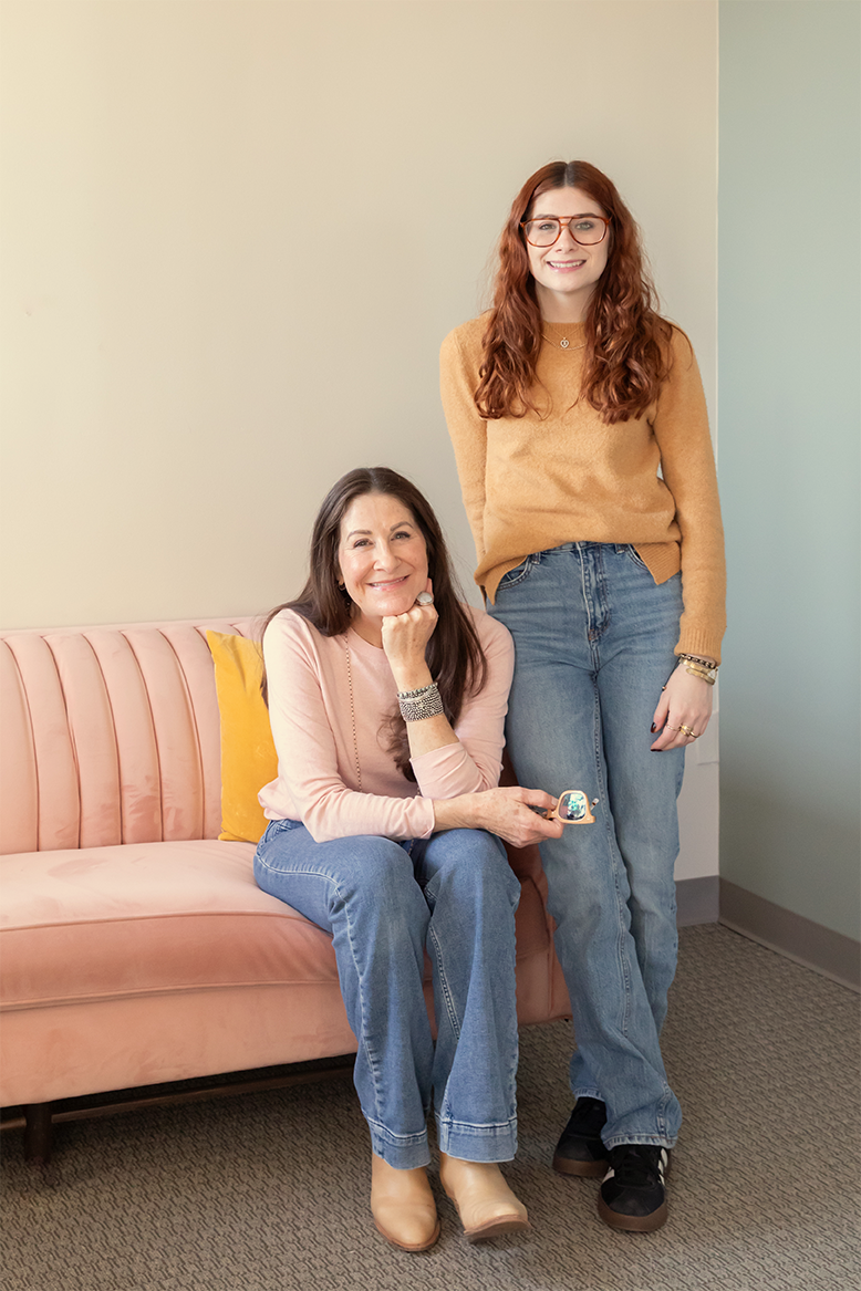 Dana Marcus, specializing in ADHD, sits on the pink velvet couch in their office in Philadelphia, while Molly Marcus, specializing in DBT & Decluttering, stands next to her.