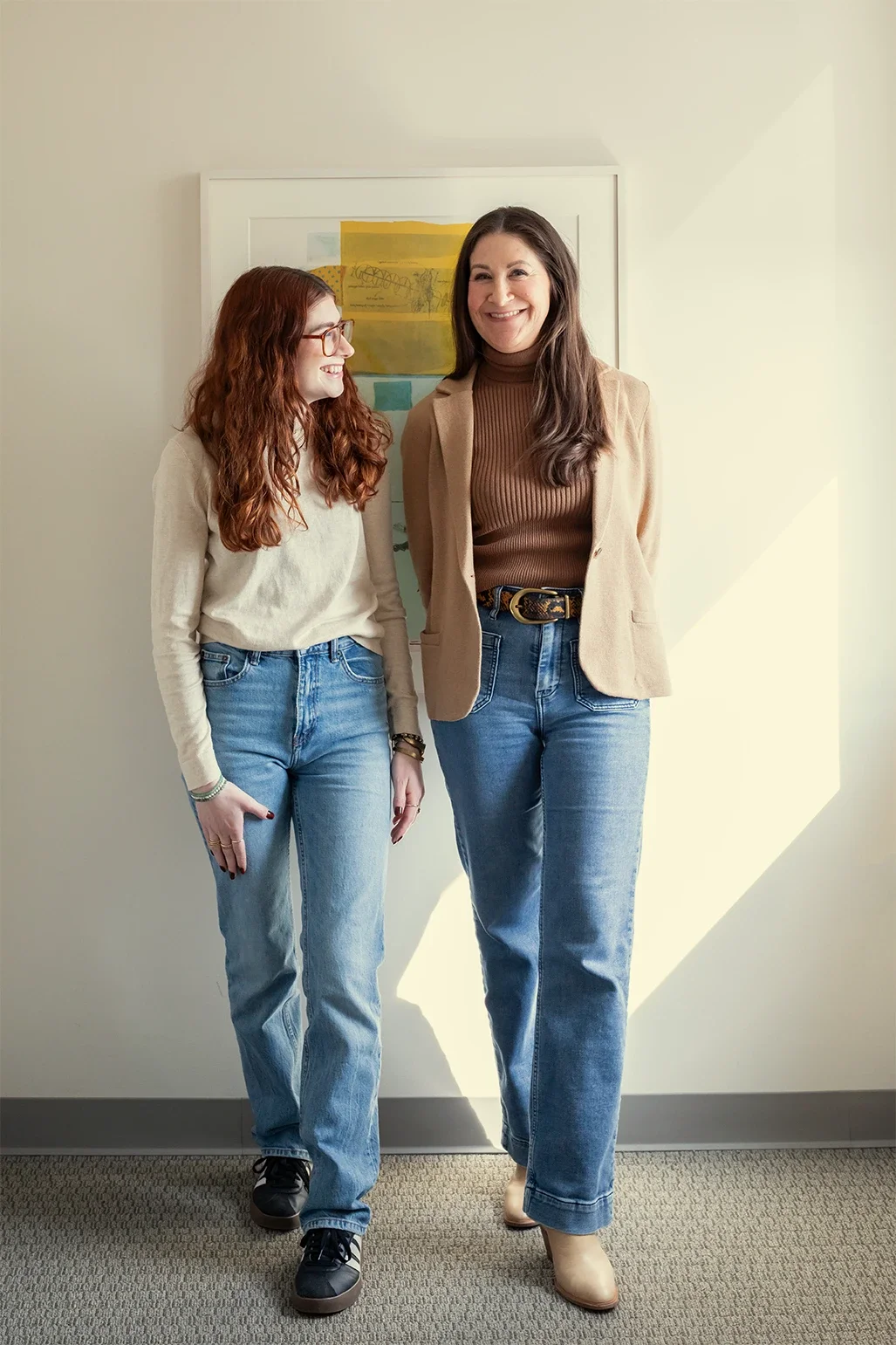 Molly Marcus, specializing in DBT & Decluttering, and Dana Marcus, specializing in ADHD, standing near a framed painting in their therapy + coaching office in Philadelphia.  Dana is smiling & looking forward; Molly is smiling & looking at Dana.