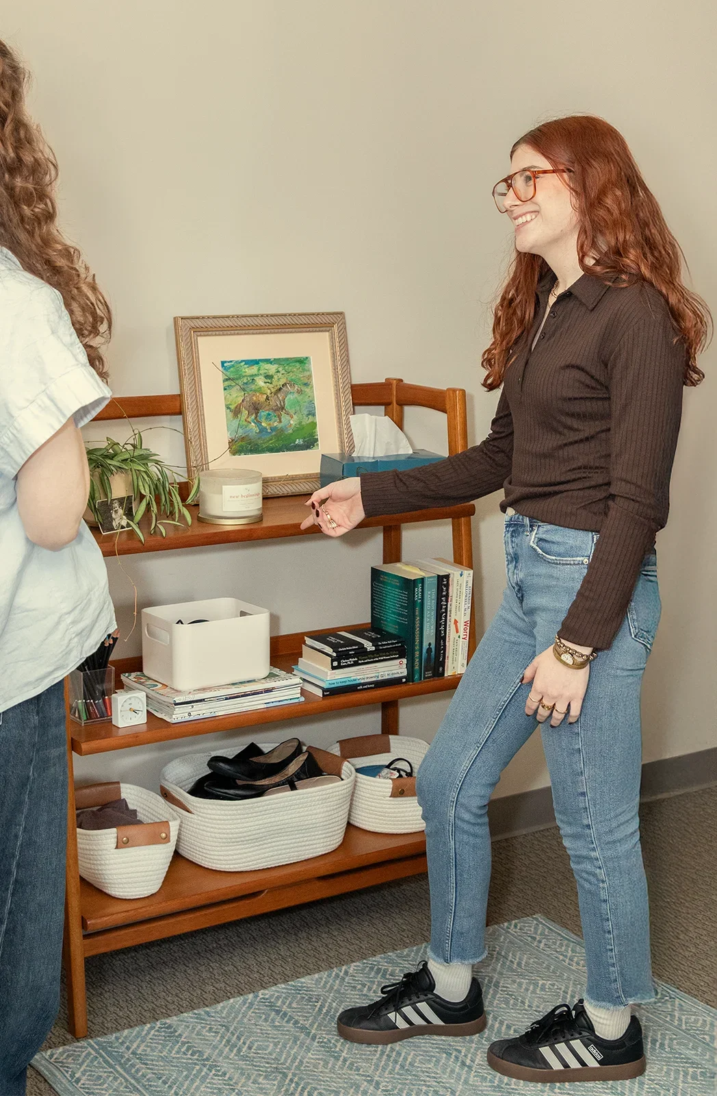 Two women having a conversation in a room with a wooden bookshelf. The woman on the right is smiling and pointing towards the shelf, wearing glasses, a dark long-sleeve shirt, and jeans. The woman on the left is partially visible, wearing a light-colored shirt.