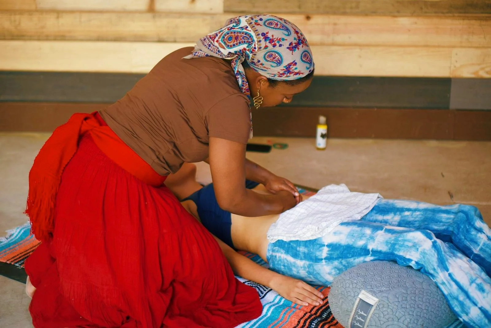 A woman providing a massage or acupuncture to a man lying face down on a mat, with a patterned laundry or cloth covering his lower body, in a room with wooden walls.