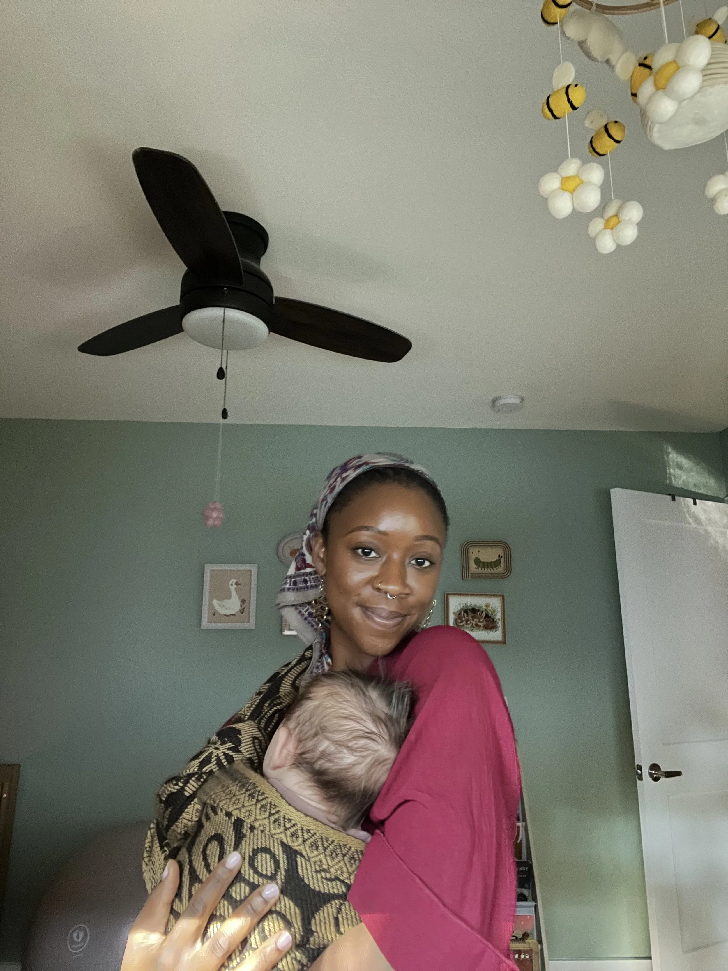 A young woman with a headscarf and nose ring holding a sleeping baby wrapped in a patterned blanket in a room with a green wall, framed wall art, a ceiling fan, and decorative hanging items.