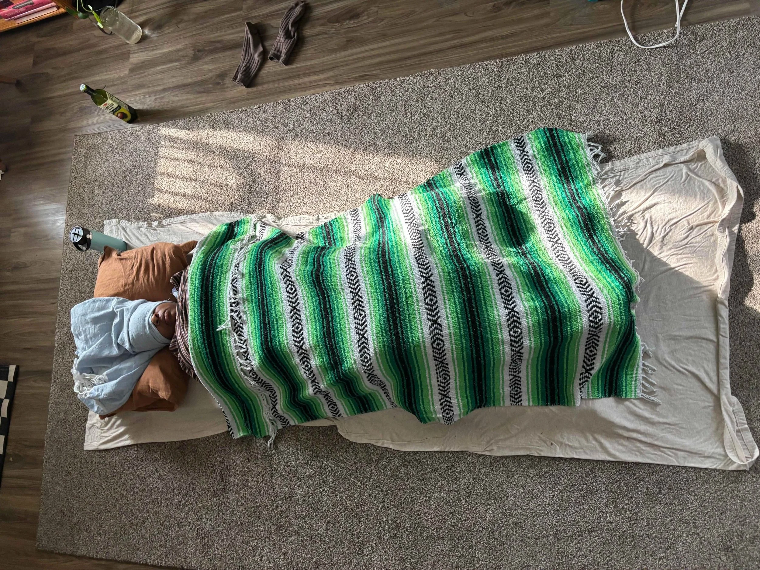 Person lying on their back on a mattress with their head on a pillow, covered with a green and white striped blanket, in a room with wooden flooring and sunlight through a window.