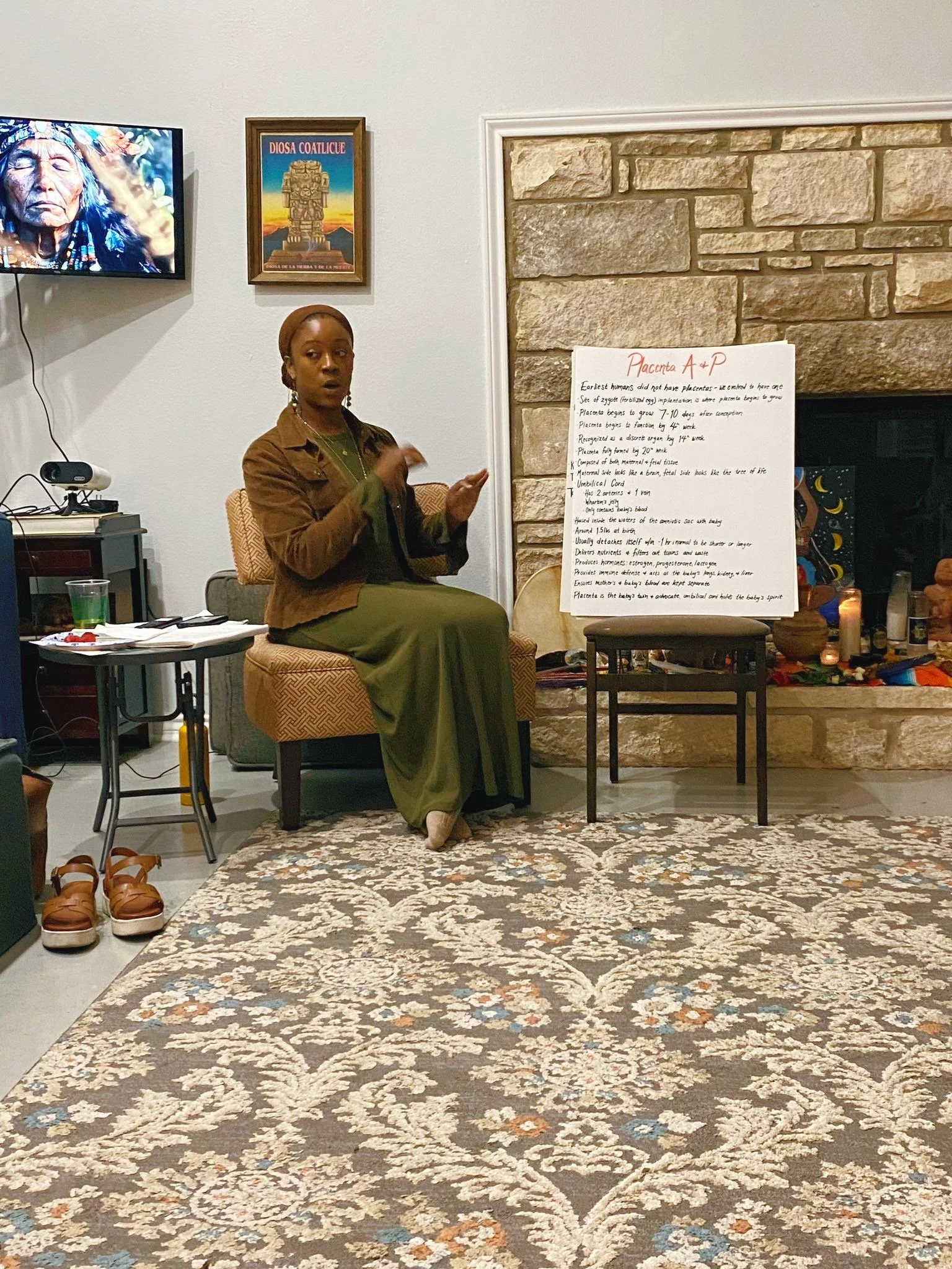 A woman sitting in a cushioned chair, giving a presentation in a living room with a stone fireplace. There is a large handwritten poster titled "Placenta A + P" next to her, along with candles, decorations, and a small table with items on it.
