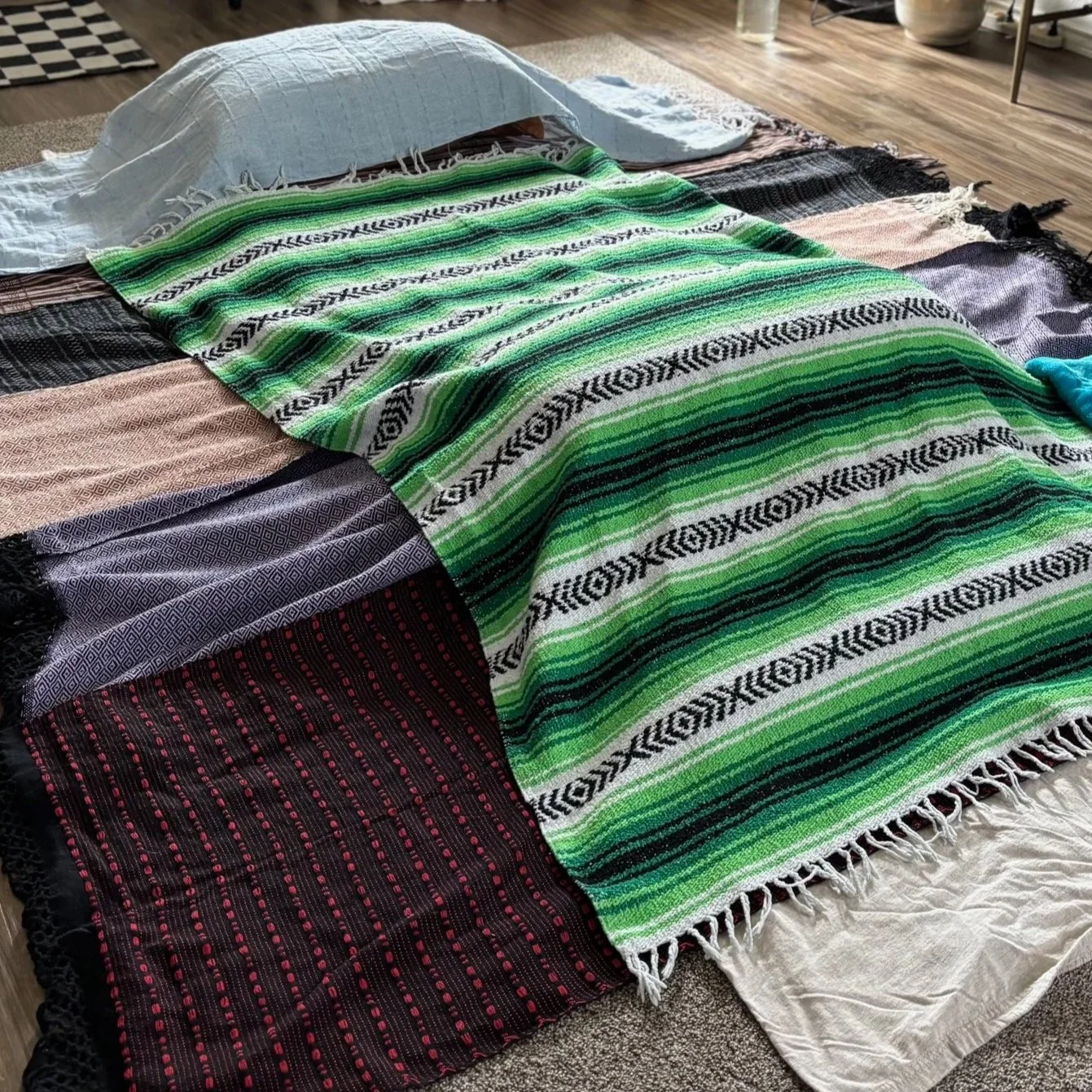 A bed with a white pillow and a colorful striped blanket on a multicolored quilt, on a wooden floor, with a glass of water and a bowl nearby.