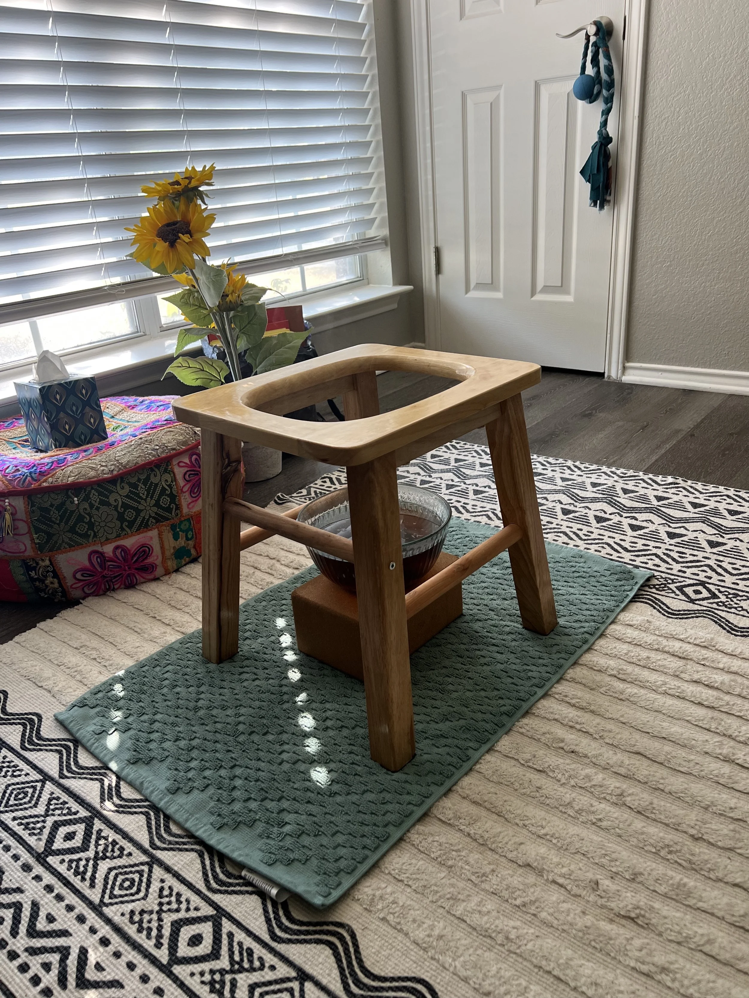 A wooden high chair with no seat on it, placed on a green textured mat. The high chair has a bowl of dark liquid or food underneath it. In the background, there is a colorful quilted cushion with a box of tissues on top, a sunflower decoration, and a window with blinds. A white door with a blue and teal tie-dye ornament hangs on the doorknob.