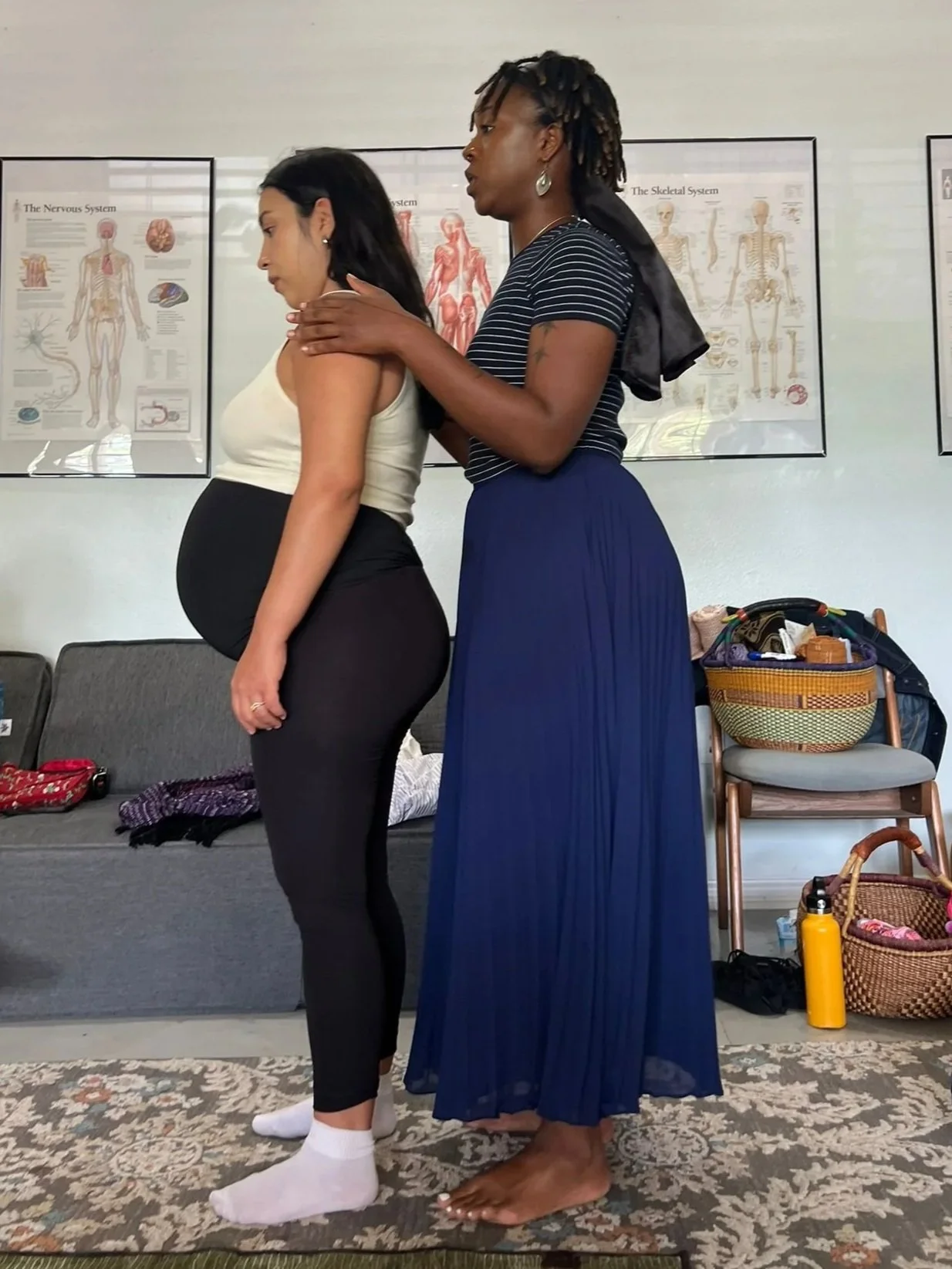 A pregnant woman stands facing forward with her eyes closed, while a woman with dreadlocks gently places her hands on the pregnant woman's shoulders in a comforting or supportive manner. They are indoors, with anatomy posters on the wall behind them and personal belongings on chairs and the floor.