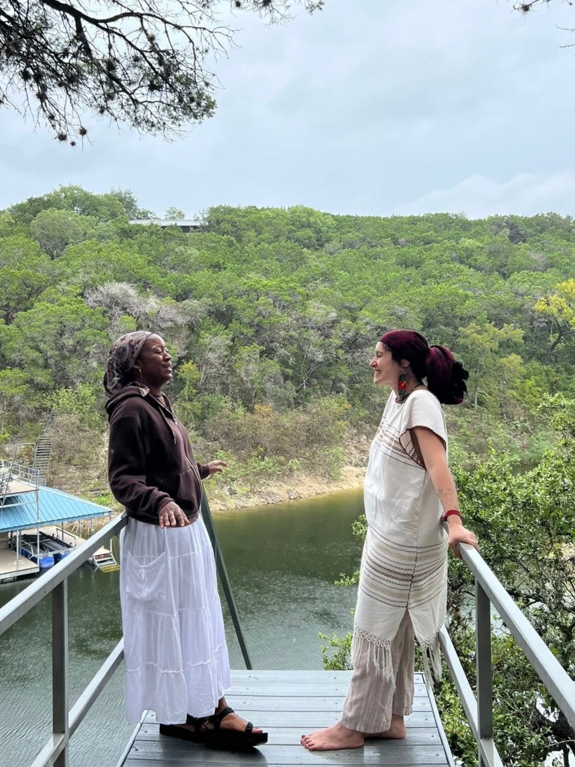 Two women standing on a wooden viewing platform over a river, smiling and talking. The woman on the left is wearing a headscarf, hoodie, and white skirt, while the woman on the right has dark hair in a bun, wearing a white dress and standing barefoot. Green trees and a cloudy sky are in the background.