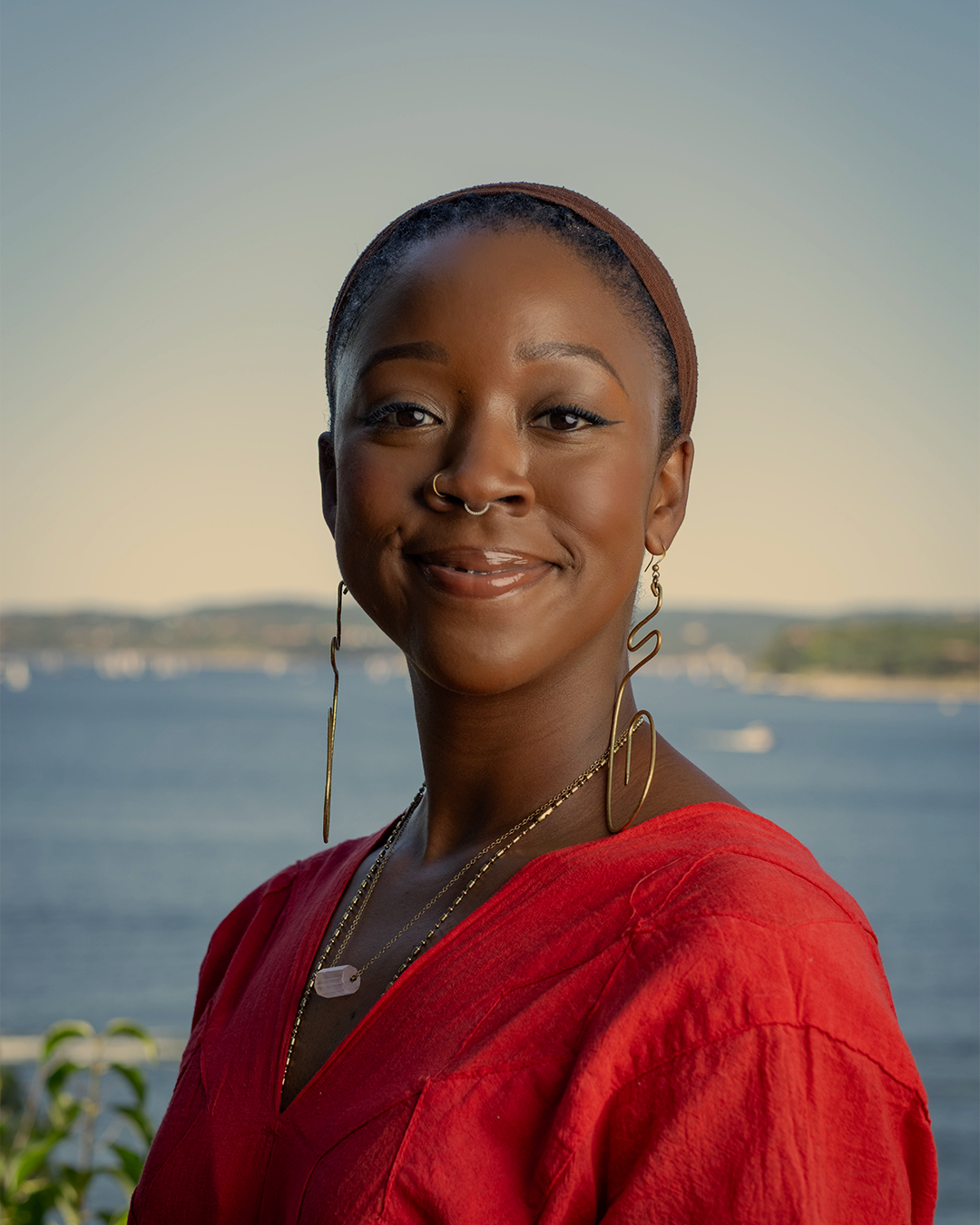 A young woman with dark skin, short hair, and earrings stands outdoors near water. She is smiling and wearing a red top and gold jewelry, with a scenic body of water and distant landscape in the background.