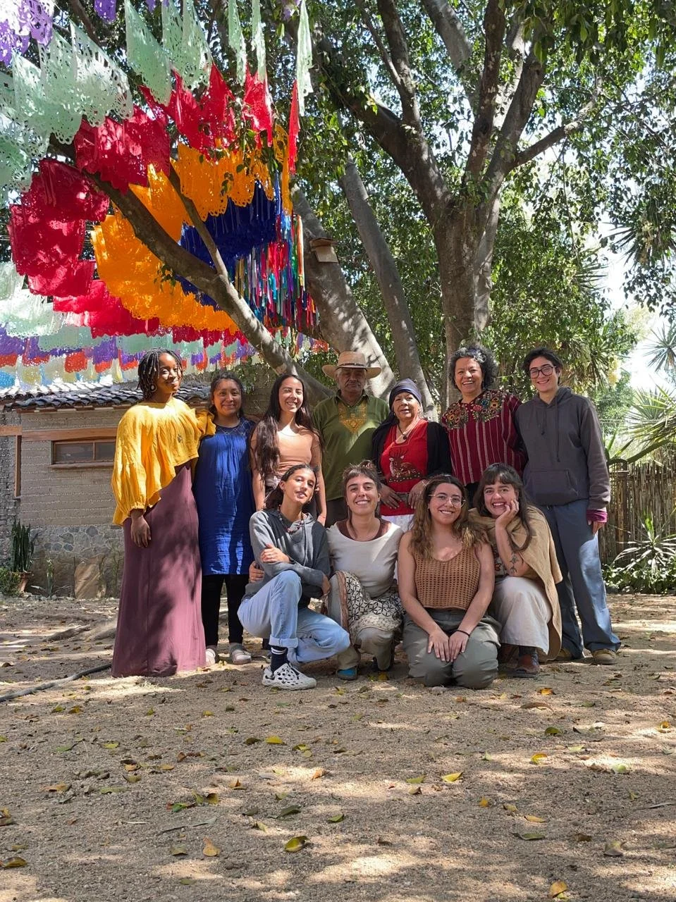 A group of ten people, diverse in age and ethnicity, pose outdoors in front of a large tree and colorful papel picado banners hanging from the branches. The group is smiling and appears to be enjoying a festive gathering.
