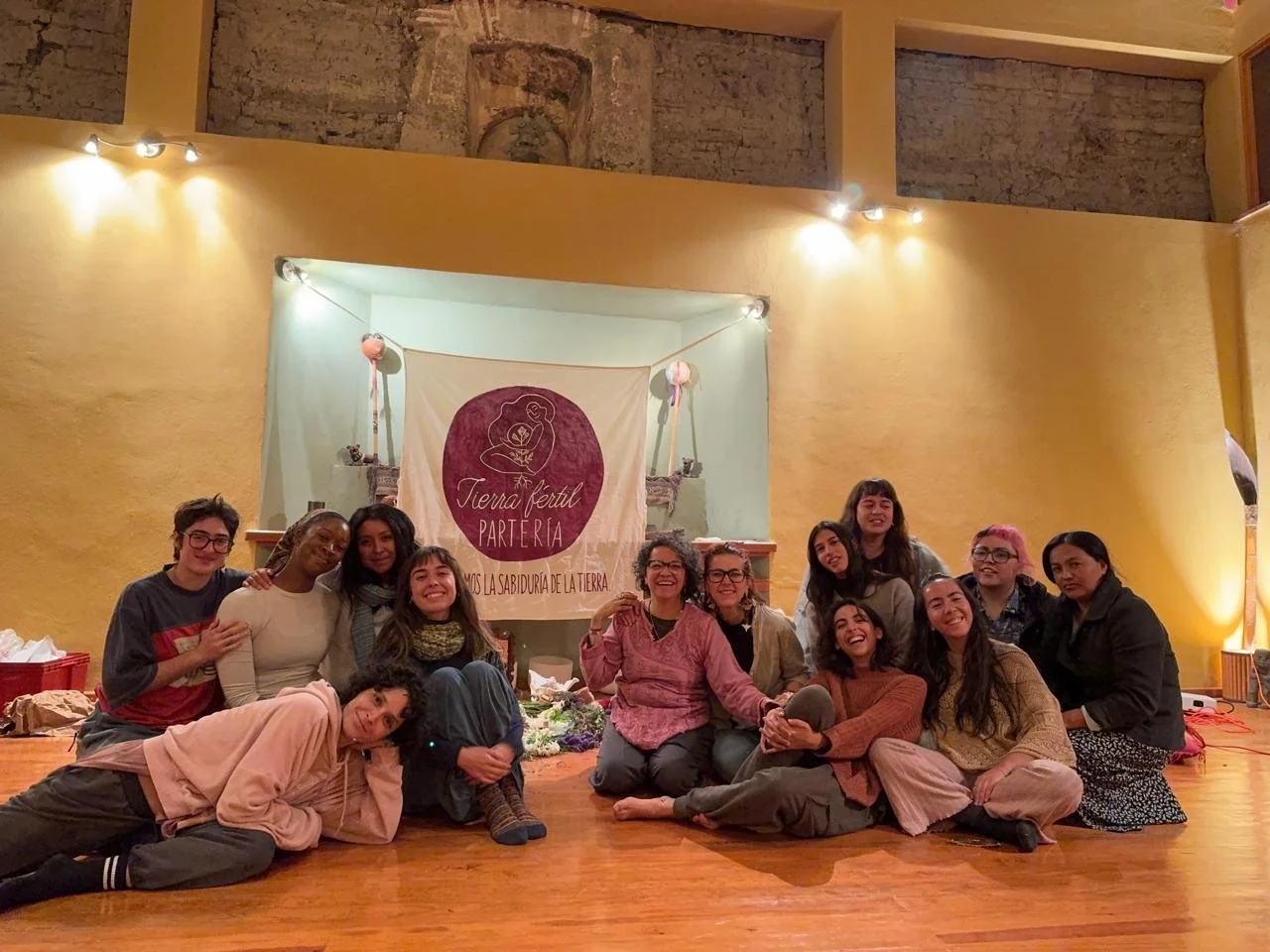 Group of women sitting and lying on the wooden floor in a cozy indoor space, smiling and posing for the photo, with a yellow wall and a banner reading 'Tierra Fértil Parte Ría' in the background.