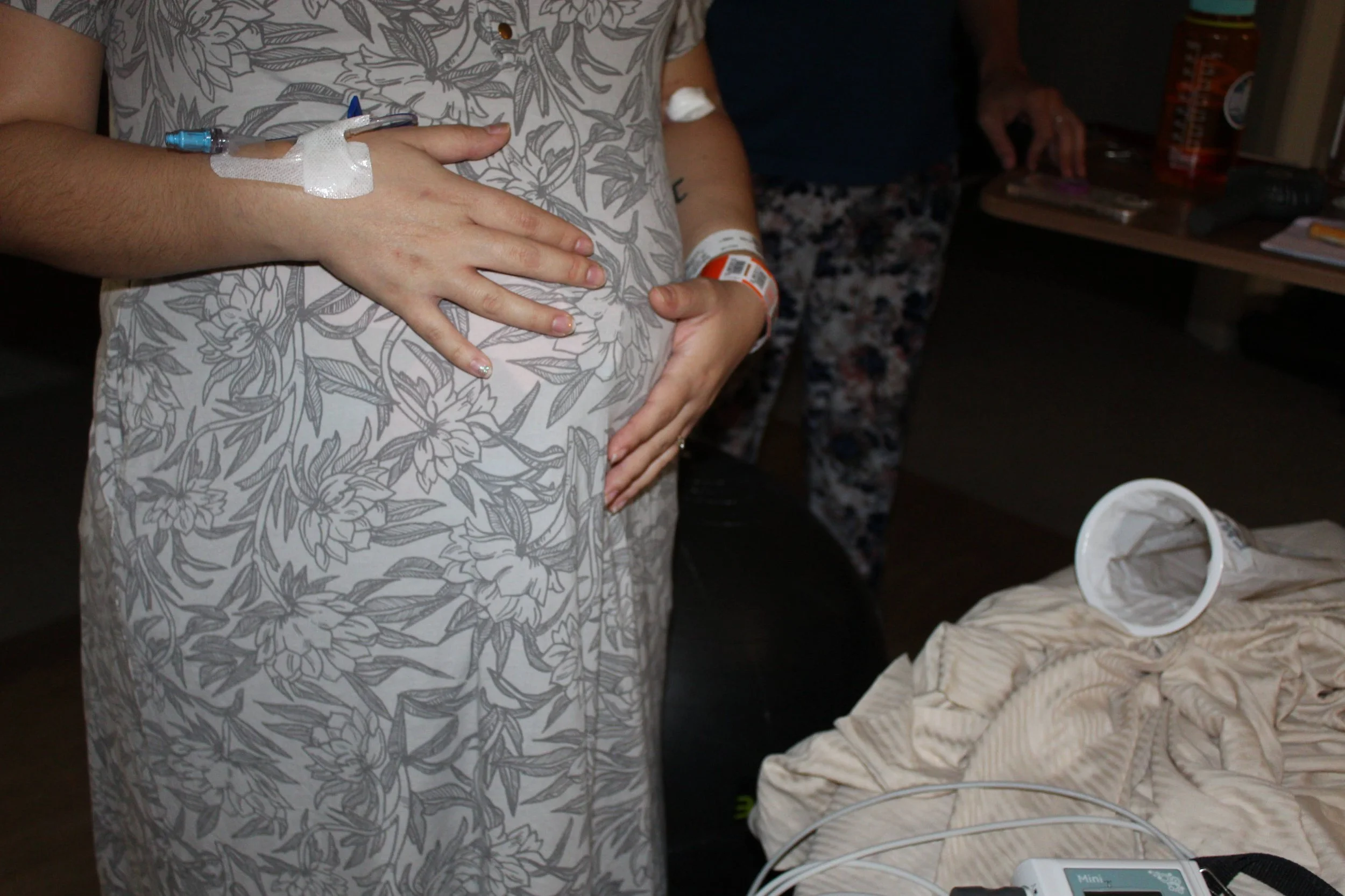 A person with hospital wristbands and intravenous (IV) line on their hand, standing next to someone else in a hospital room. The person is wearing floral-patterned clothing and appears to be pregnant.