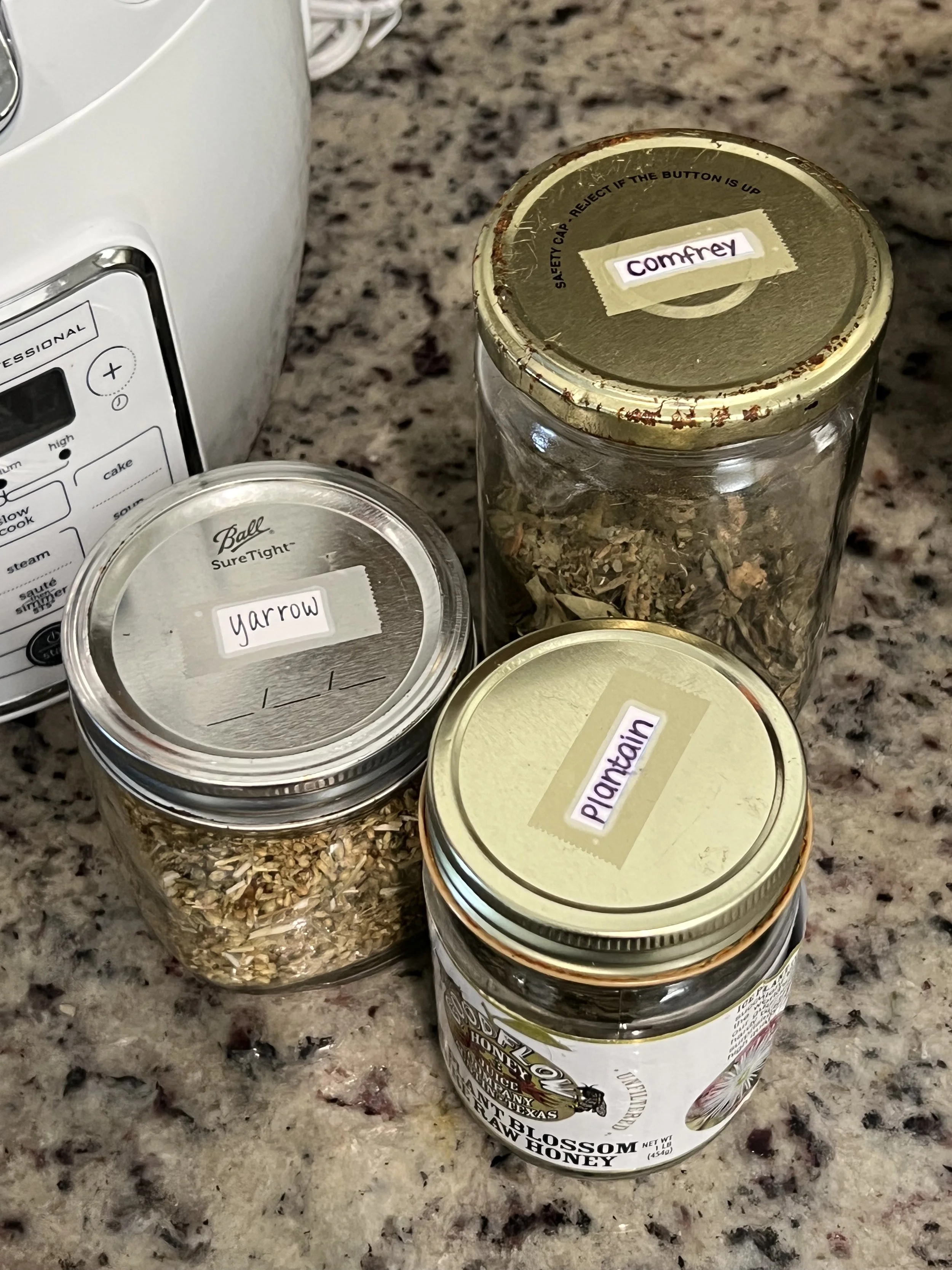 Four glass jars with metal lids, each labeled with different herbs and spices: 'comfrey,' 'yarrow,' 'plantain,' and 'wildflower honey,' placed on a granite countertop next to a microwave oven.