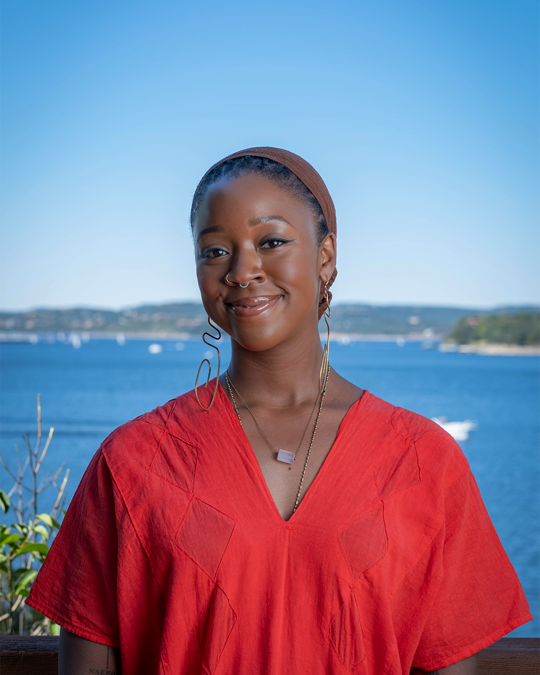 A smiling woman with dark skin wearing a red top, jewelry, and a brown headscarf standing outdoors against a lake and landscape background.