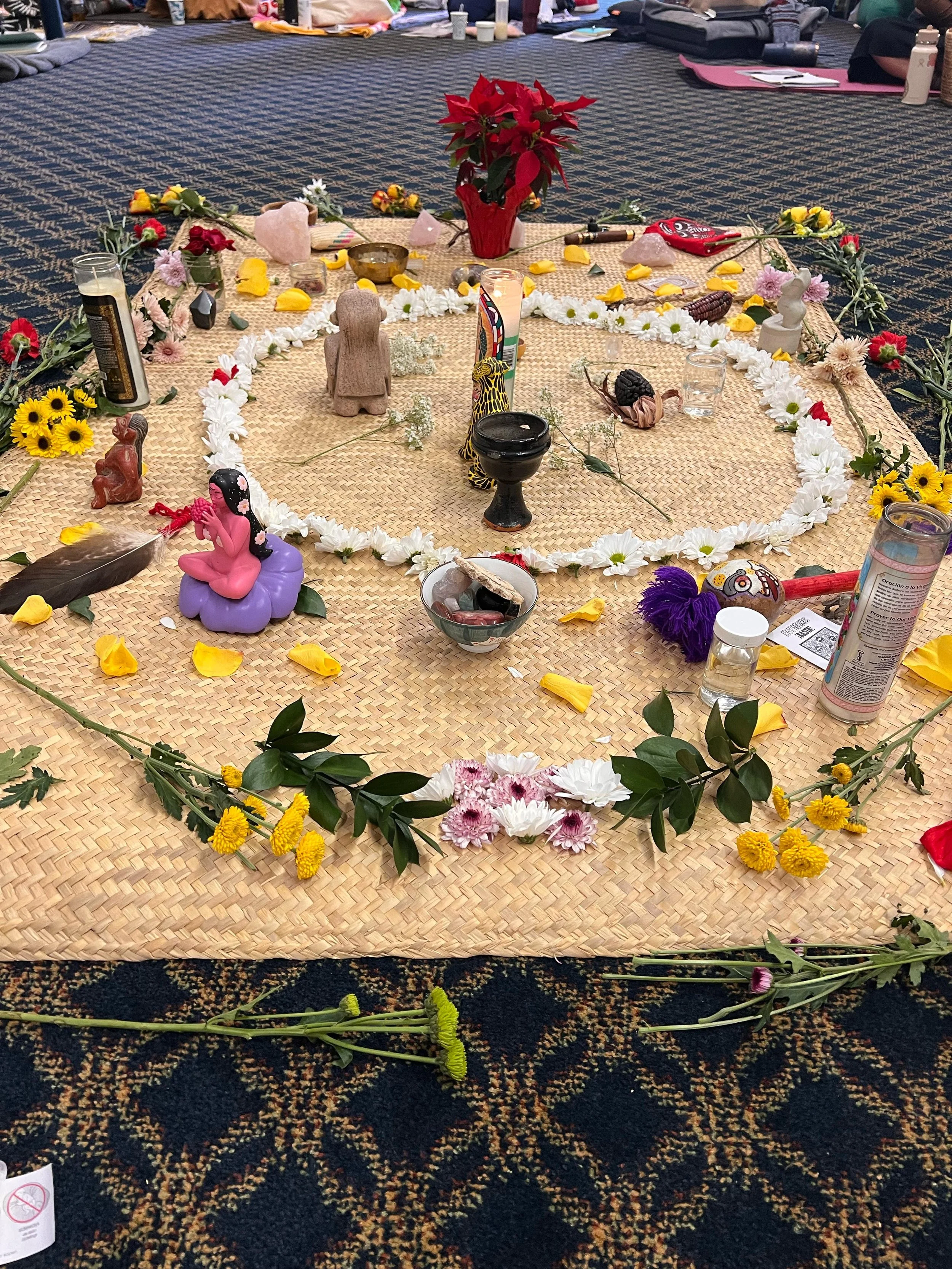 A circular arrangement of flowers, candles, and spiritual objects on a woven mat, with people sitting on yoga mats in the background.