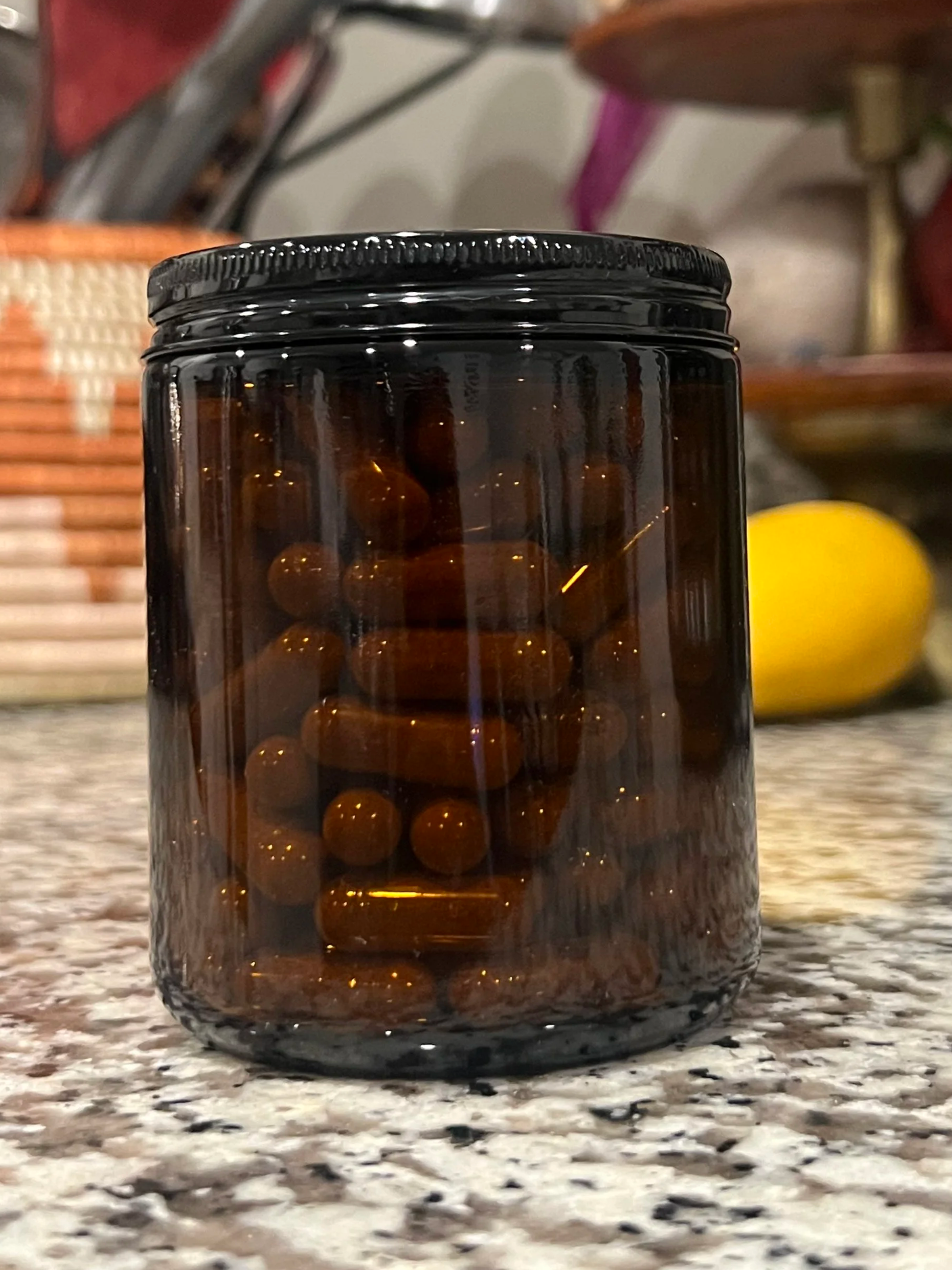 A black glass jar filled with brown capsules on a speckled countertop, with a blurred background including a yellow lemon.