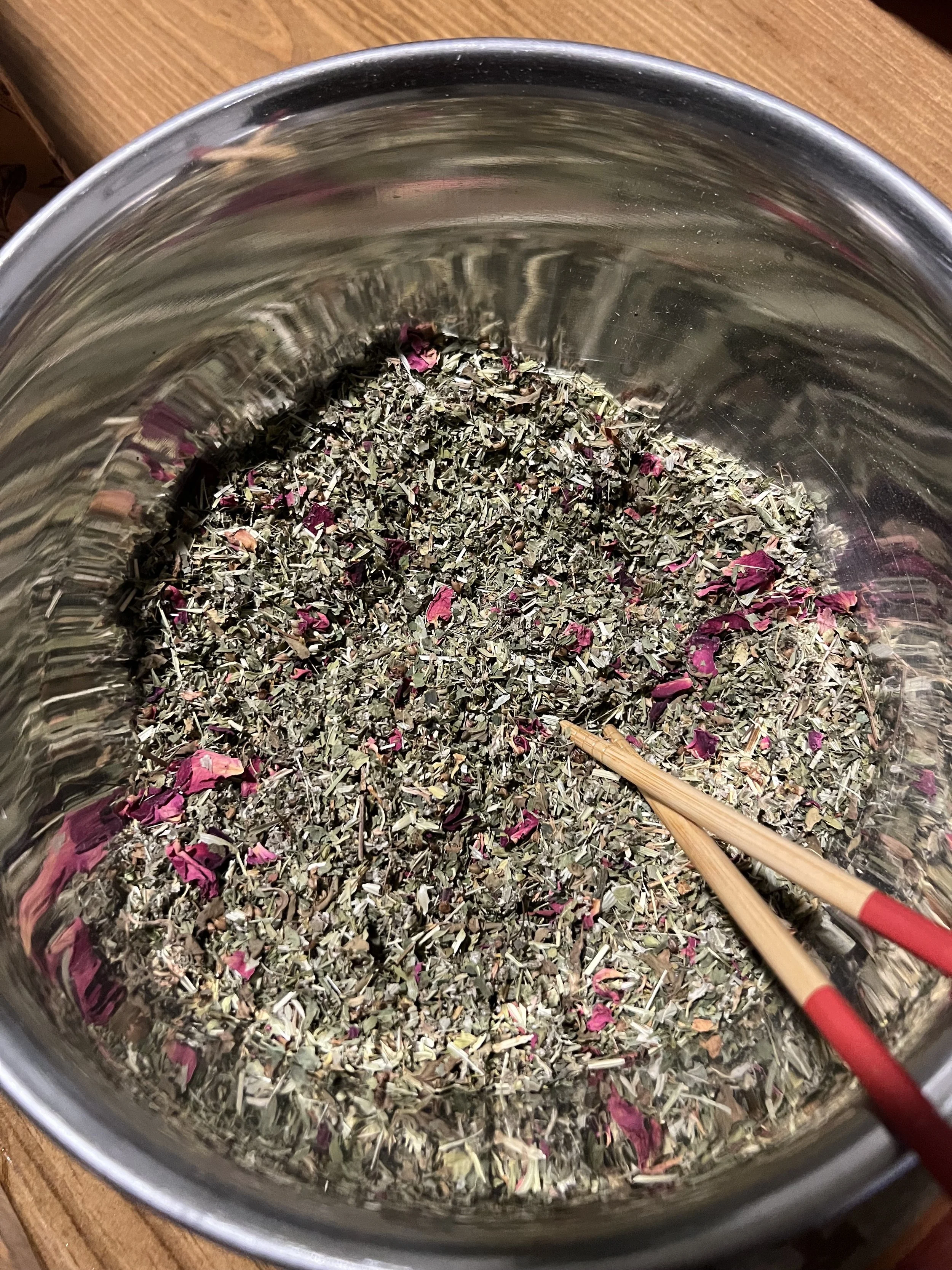 A large metal container filled with dry herbal mixture and rose petals, with a wooden spoon resting inside.