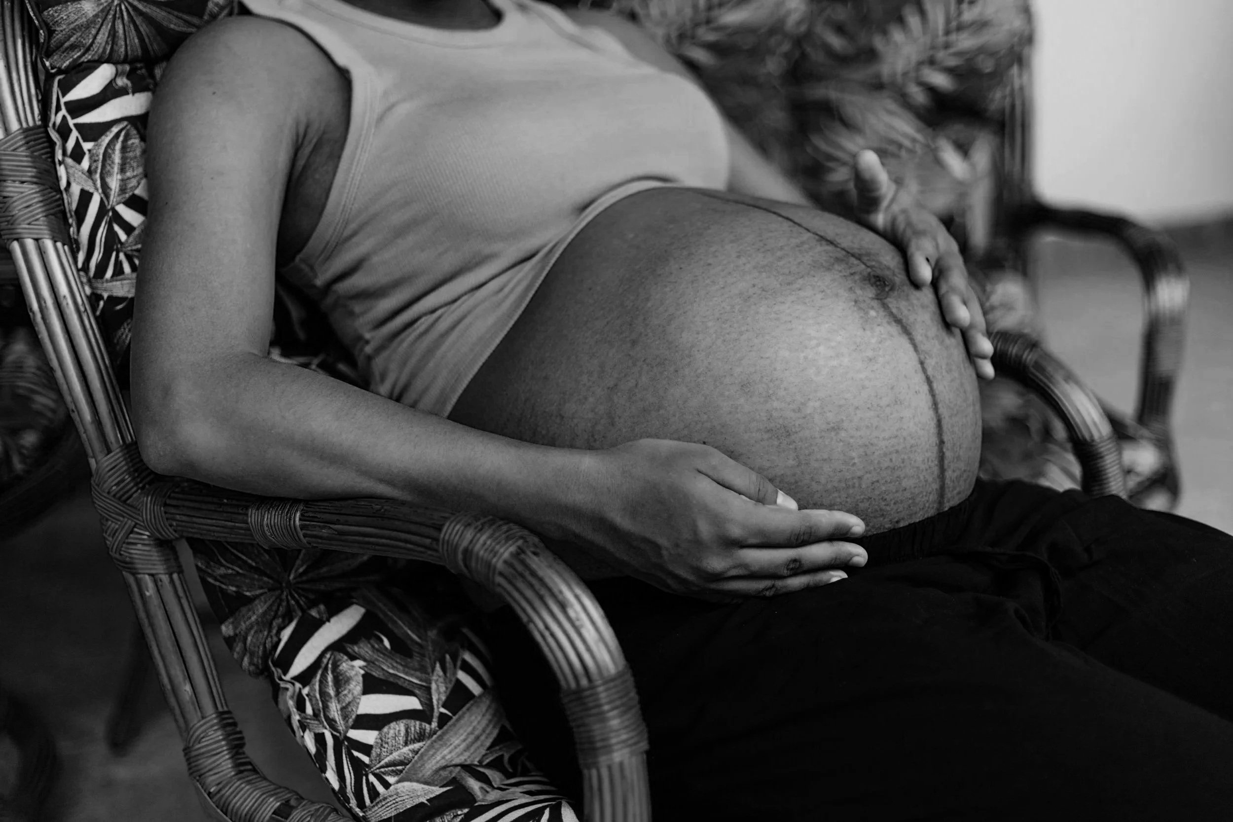 A person with a large pregnant belly sitting on a chair, resting their hand on their abdomen. The image is in black and white, and the person is wearing a sleeveless shirt.