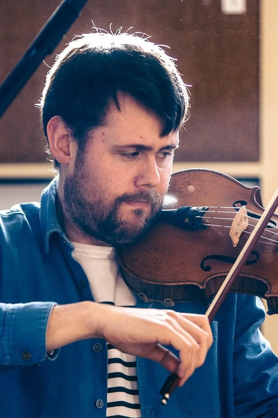 A man with dark hair and a beard playing the violin.