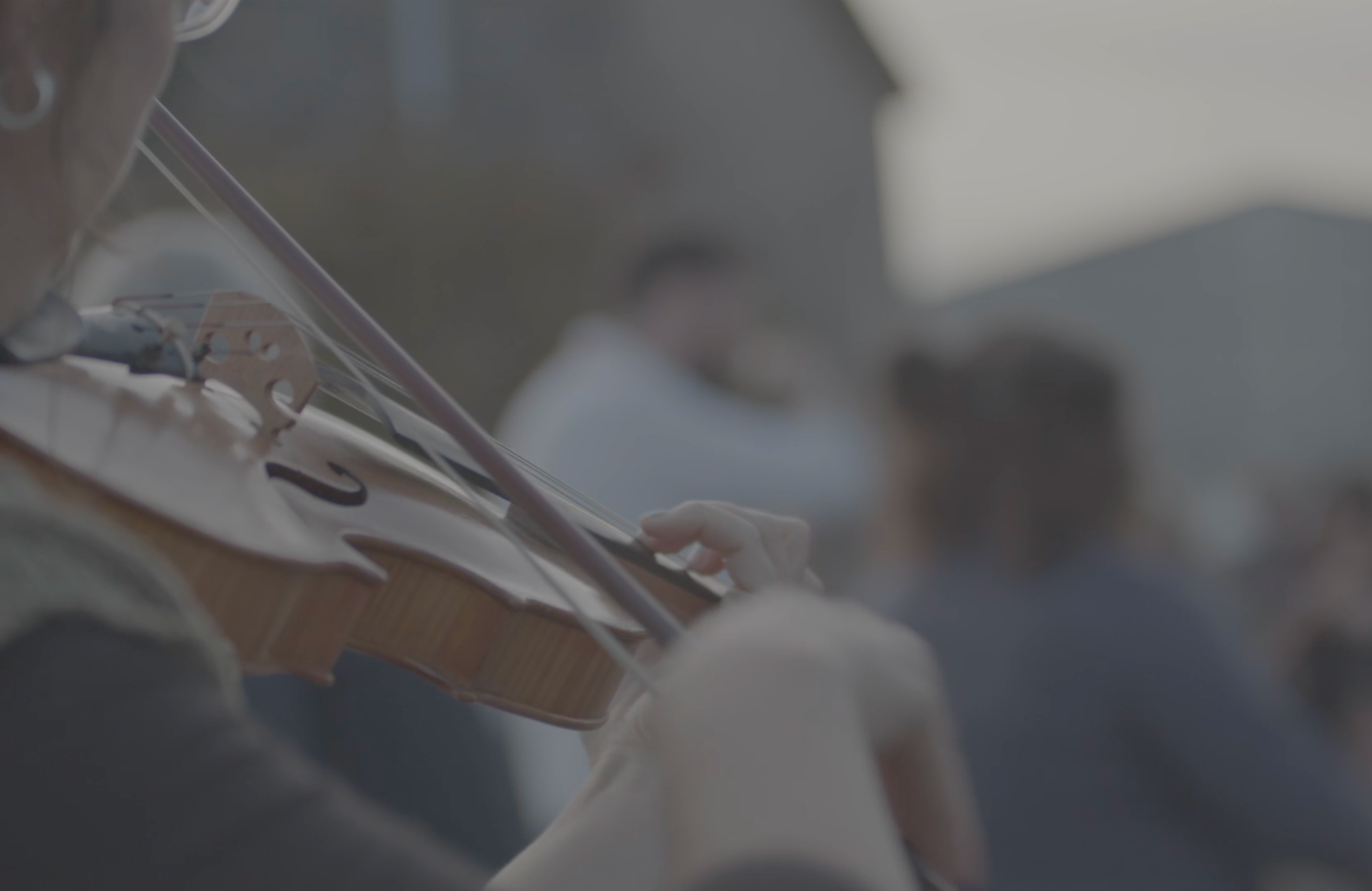 A person playing a violin outdoors with a blurred woman in the background.