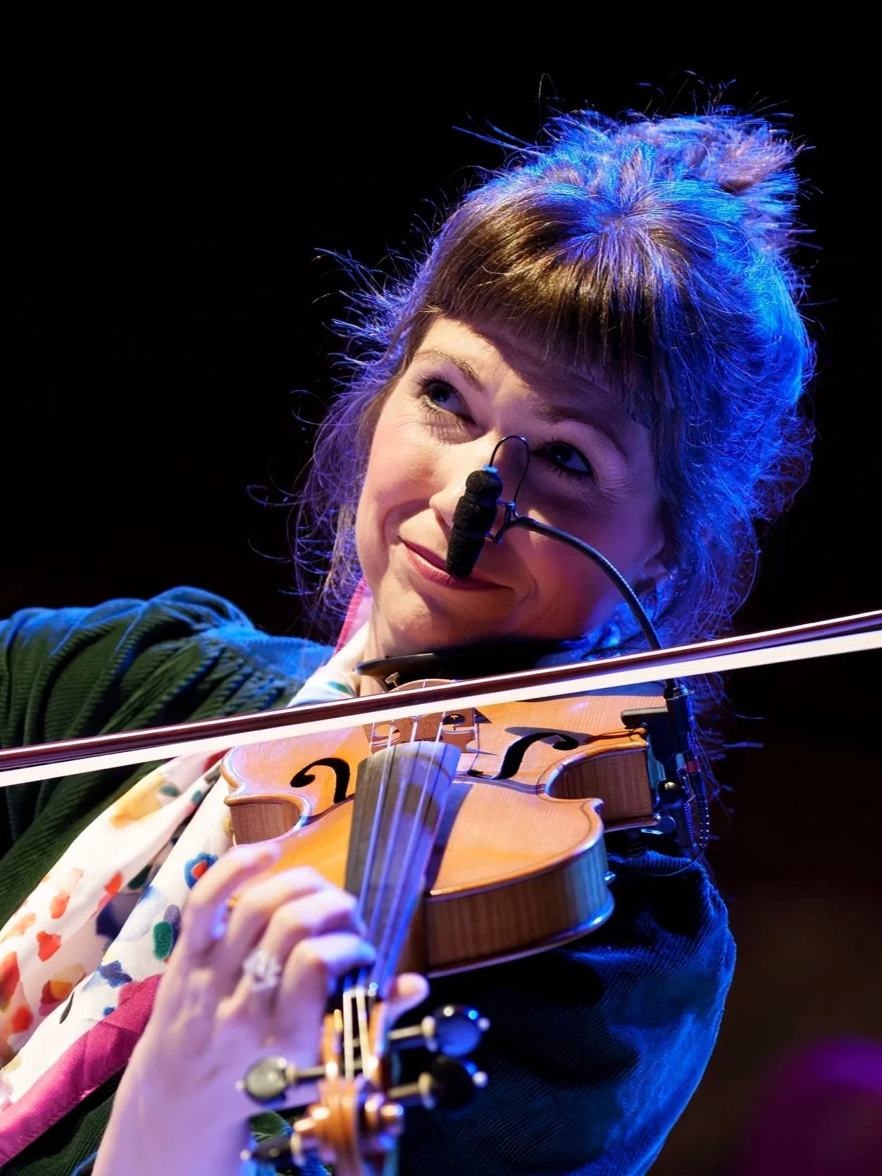 A woman playing the violin on stage, wearing a headset microphone, with blue and purple stage lighting.