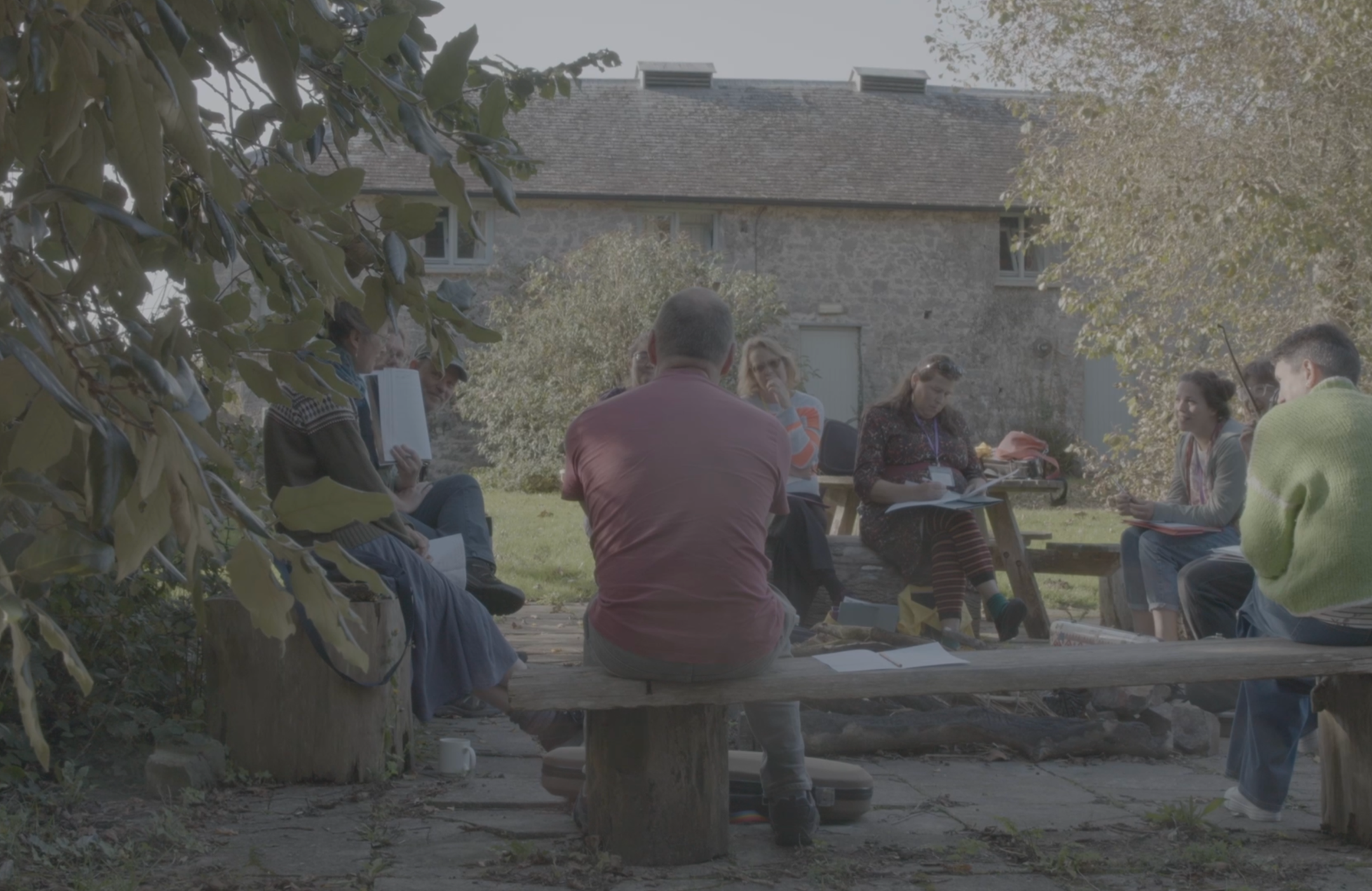 Group of people sitting outdoors in a semi-circle, engaged in a discussion or meeting, with a building and trees in the background.