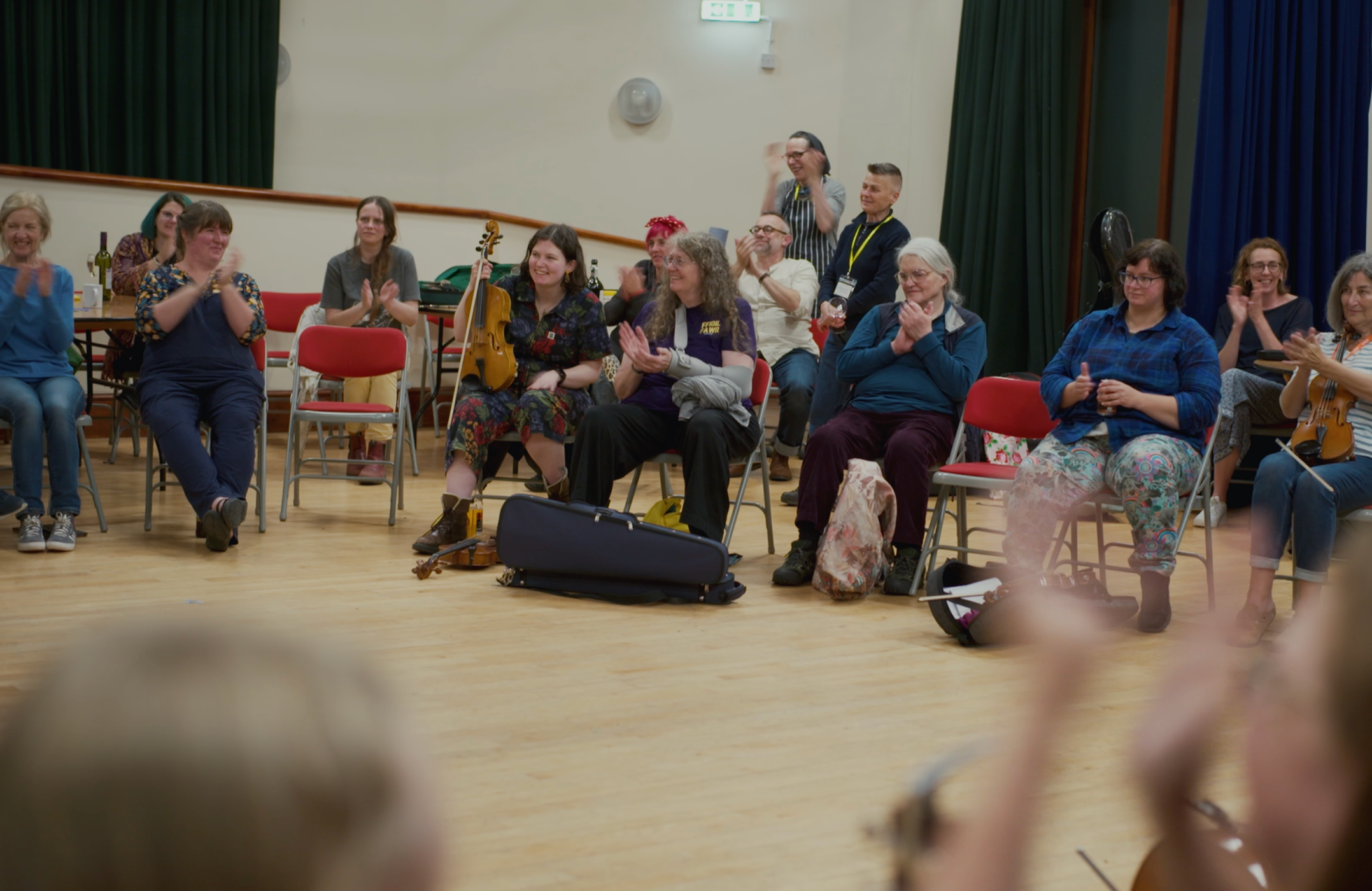 Group of people sitting and standing, clapping and smiling in a room with wooden floors and blue and green curtains.