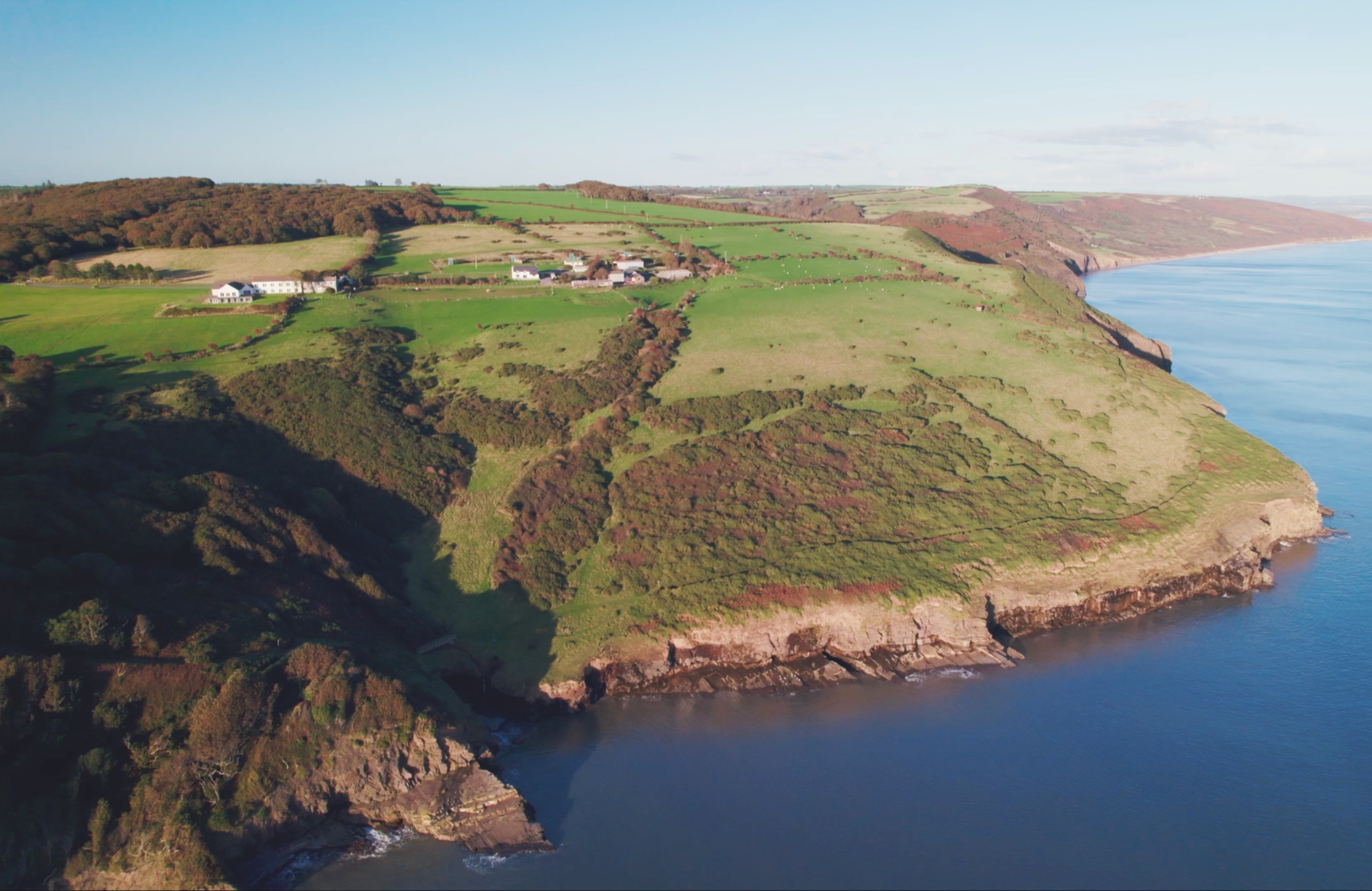 Aerial view of green coastal cliffs and farmland along the shoreline with a few white buildings, overlooking a calm blue ocean under a partly cloudy sky.