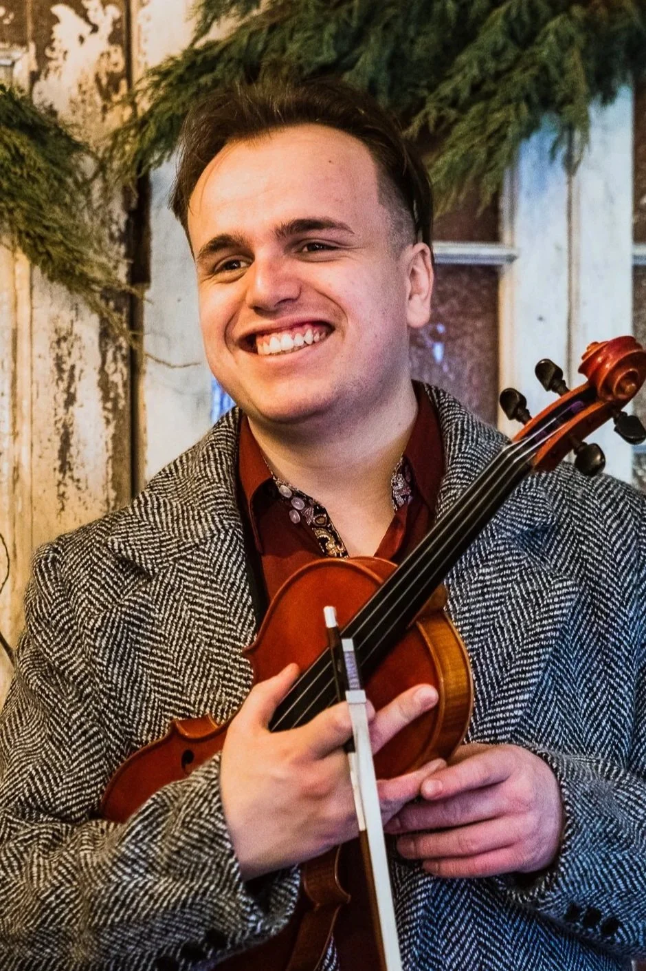 A man smiling while holding a violin in front of a rustic background decorated with greenery.
