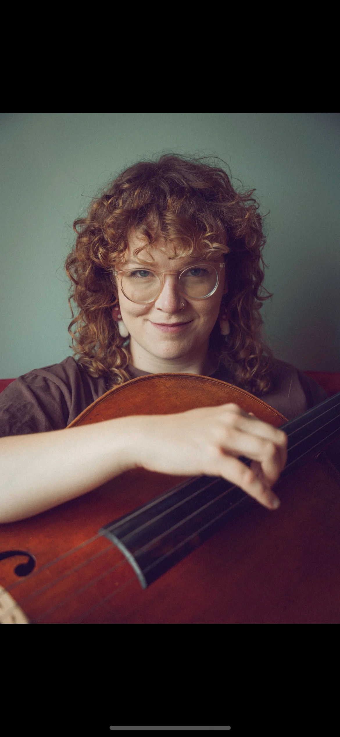 A woman with curly hair, glasses, and earrings, holding and playing an acoustic guitar.