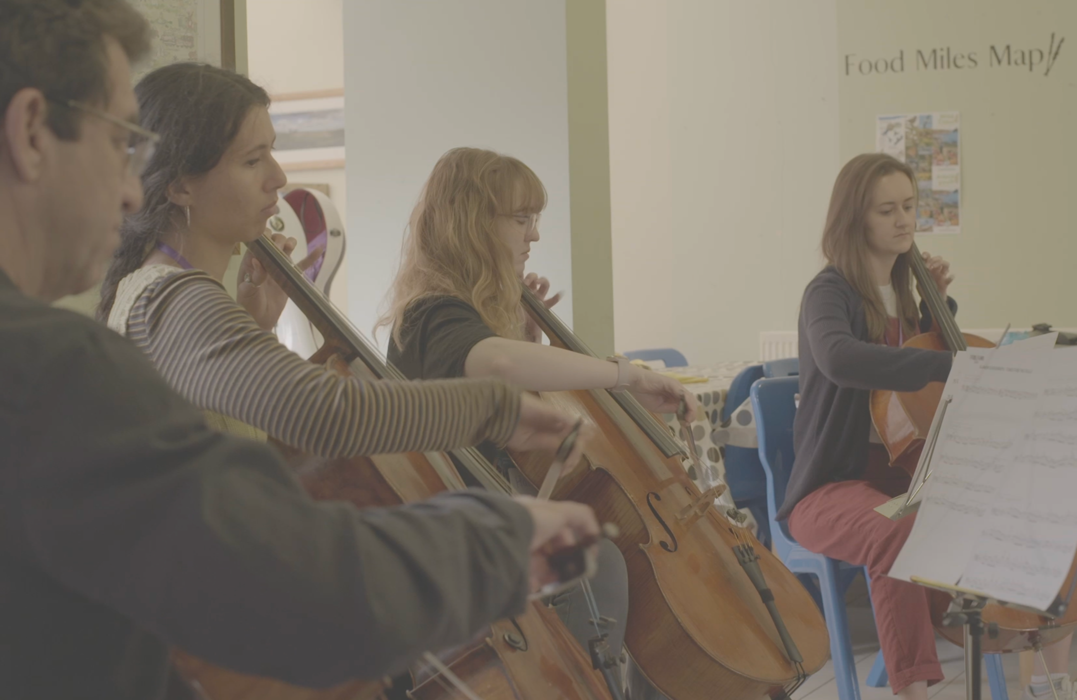 Group of four people playing string instruments in a room, including a man and three women, with sheet music on stands.
