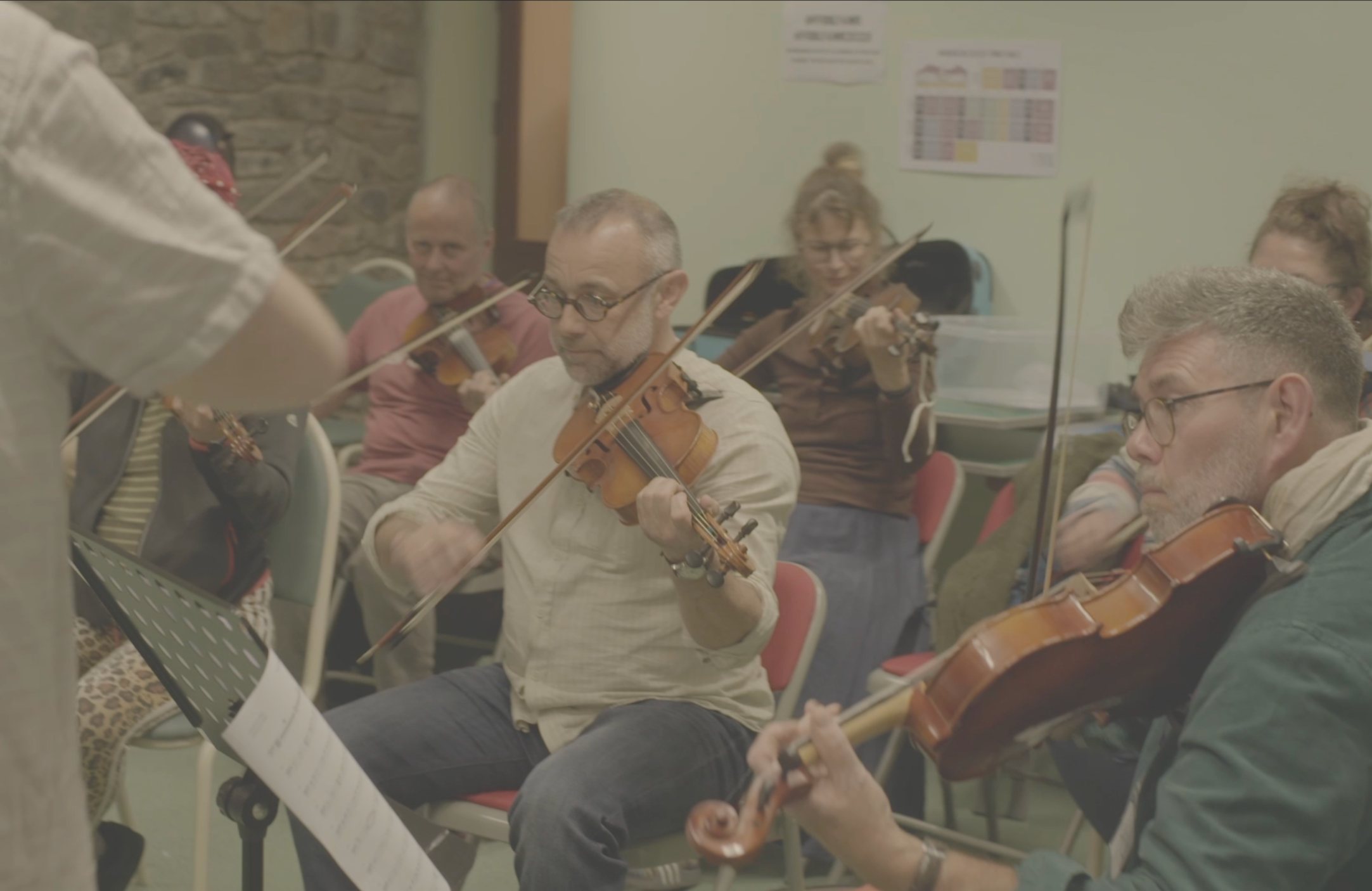 Group of musicians playing violins in a classroom or rehearsal space, seated behind a clear plastic barrier, with music sheets and posters on the wall.