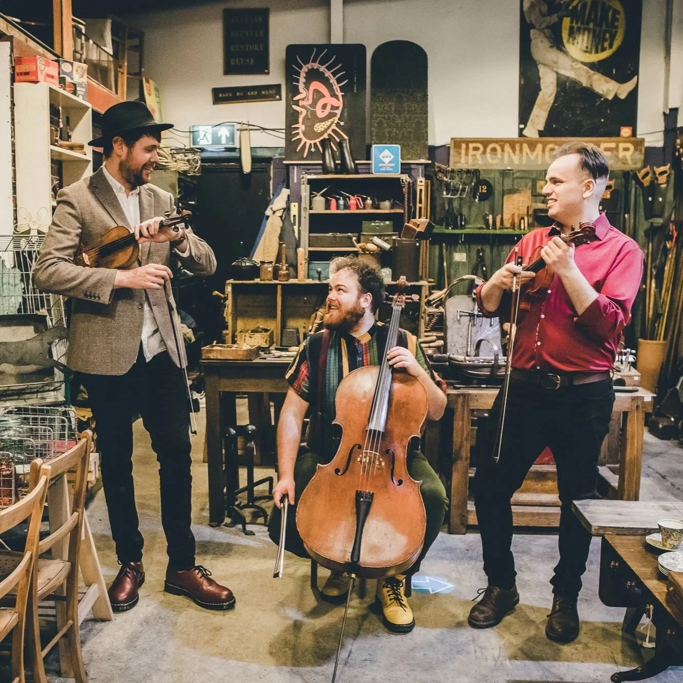 Three men with musical instruments in a workshop or store filled with tools and decor, engaging in a joyful music session.