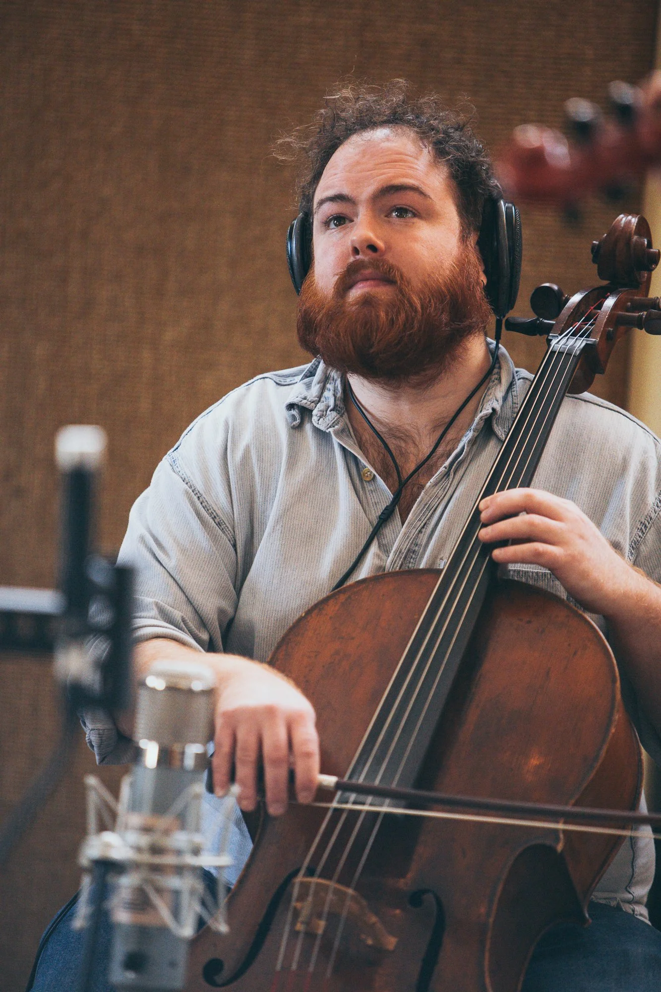 A man with a red beard and curly hair wearing headphones, playing a cello in a recording studio.