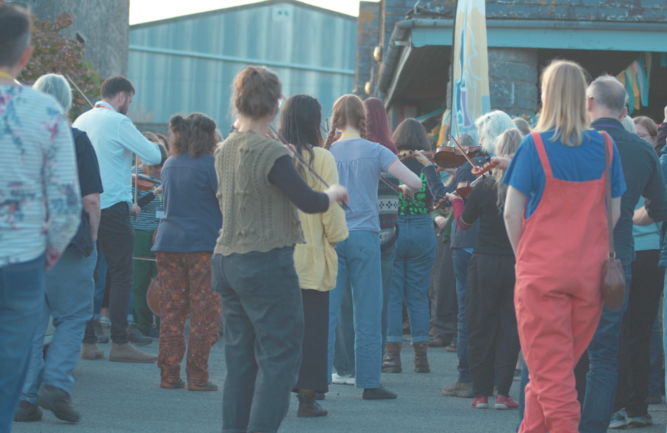 People gathered outdoors, some playing violins, in a casual, social setting