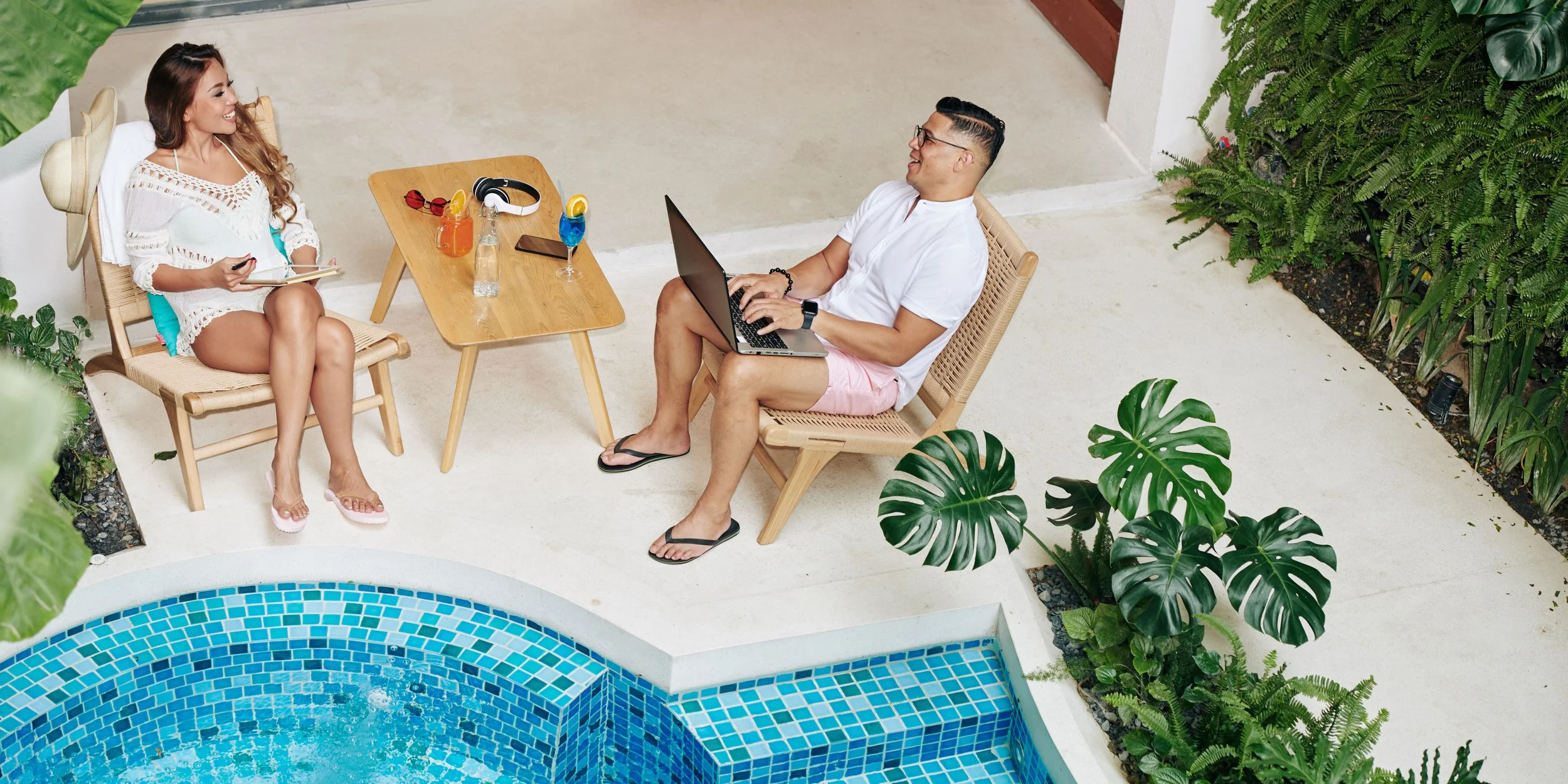 Deux personnes assises près d'une piscine, discutant et riant, avec des boissons colorées, un ordinateur portable, des écouteurs et des lunettes de soleil sur une table en bois, entourées de plantes