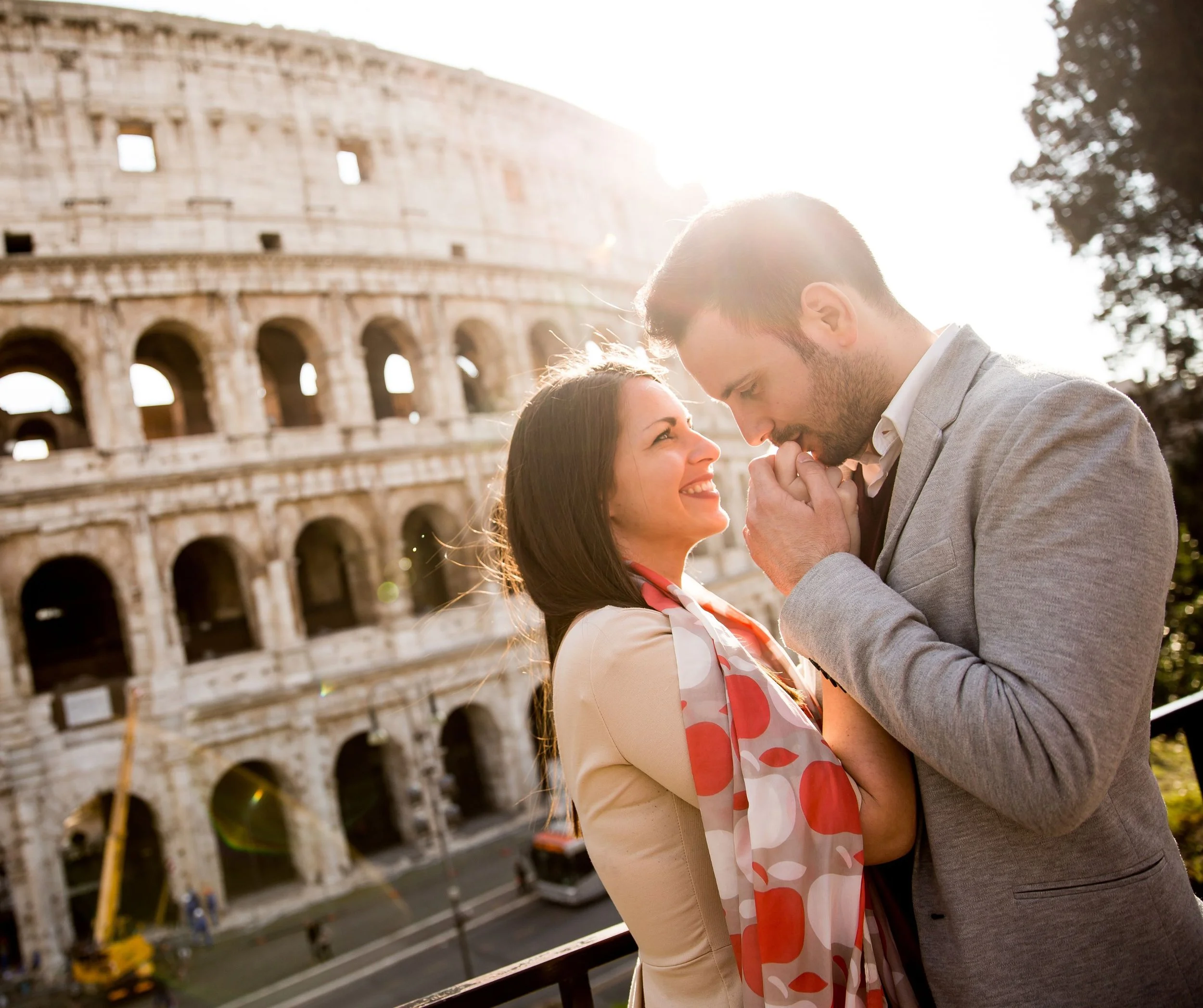 Un couple en dehors du Colisée à Rome, la femme sourit et regarde l'homme qui tient ses mains près de sa bouche, avec le soleil en arrière-plan.
