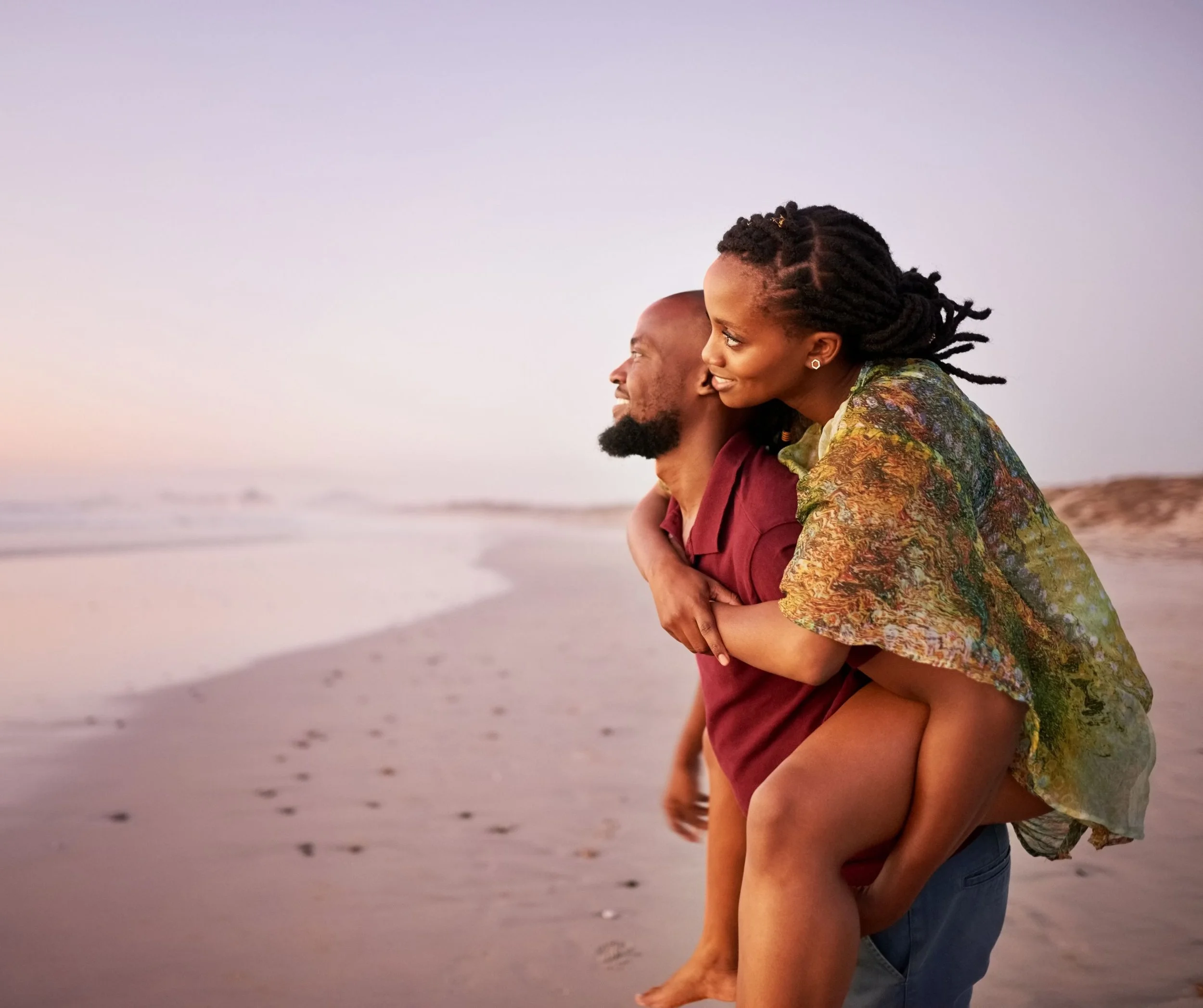 Un couple sur la plage au coucher du soleil, l'homme portant une femme sur son dos, ils sourient et profitent de la vue