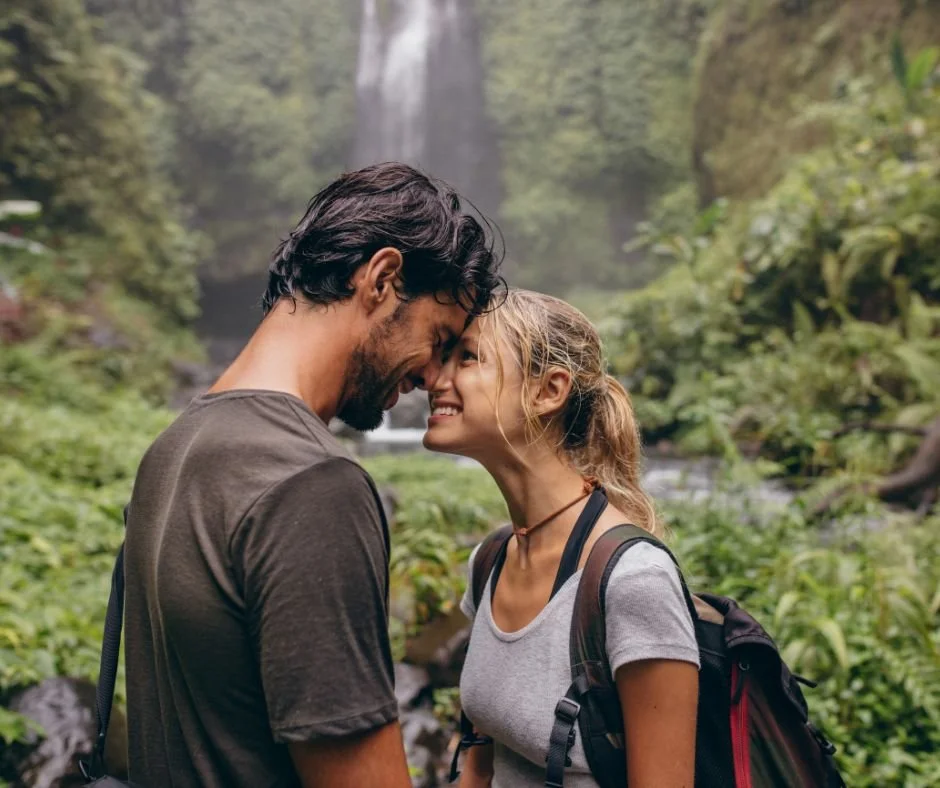 Un couple souriant se tenant face à face dans une forêt avec une cascade en arrière-plan.