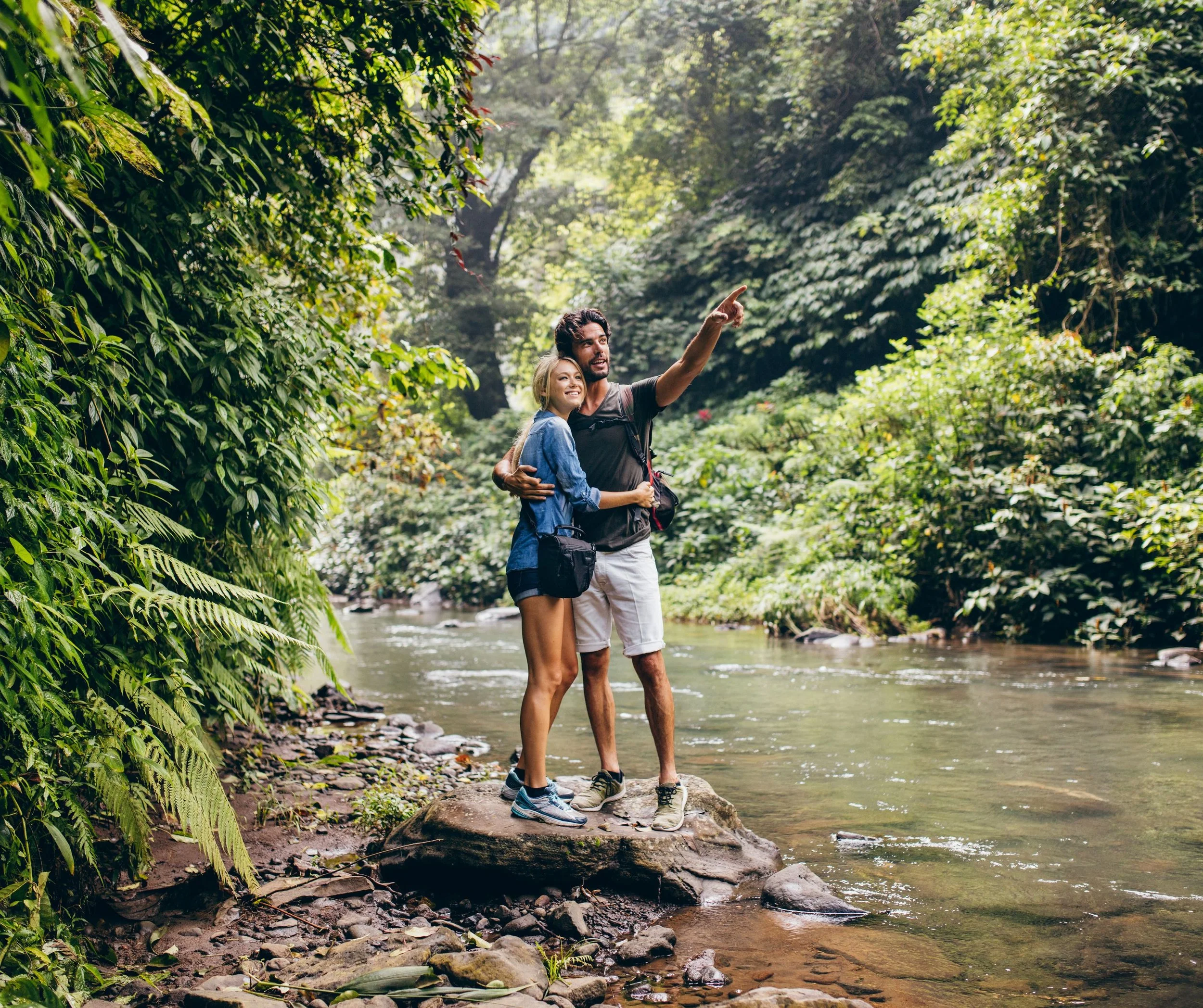 Un couple marche dans une forêt proche d'une rivière, la femme souriante écoute le homme qui lui montre quelque chose en pointant du doigt, entourés d'une végétation dense et luxuriante.