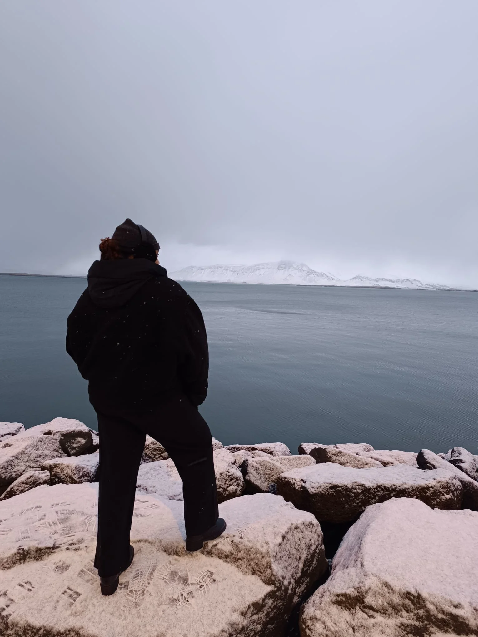 Une personne habillée chaudement regarde un lac froid entouré de montagnes enneigées, sous un ciel nuageux.